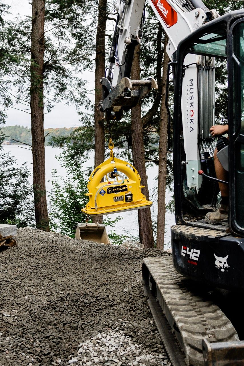 Bobcat E42 compact excavator suspending yellow MQUIP stone vacuum lifter near lake in wooded setting.