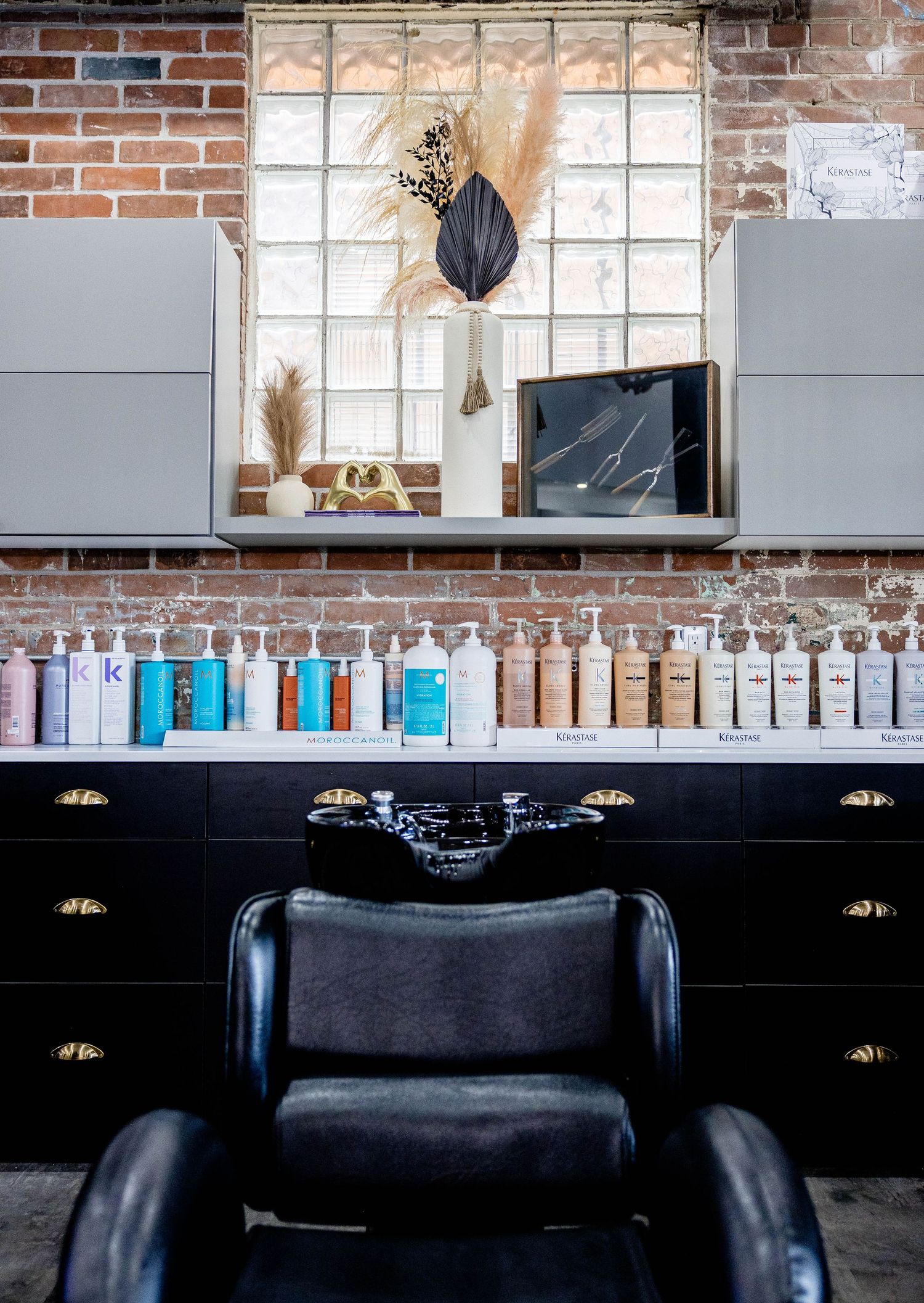 Modern hair salon station with black leather chair, product display shelf, exposed brick wall, and decorative mirror.