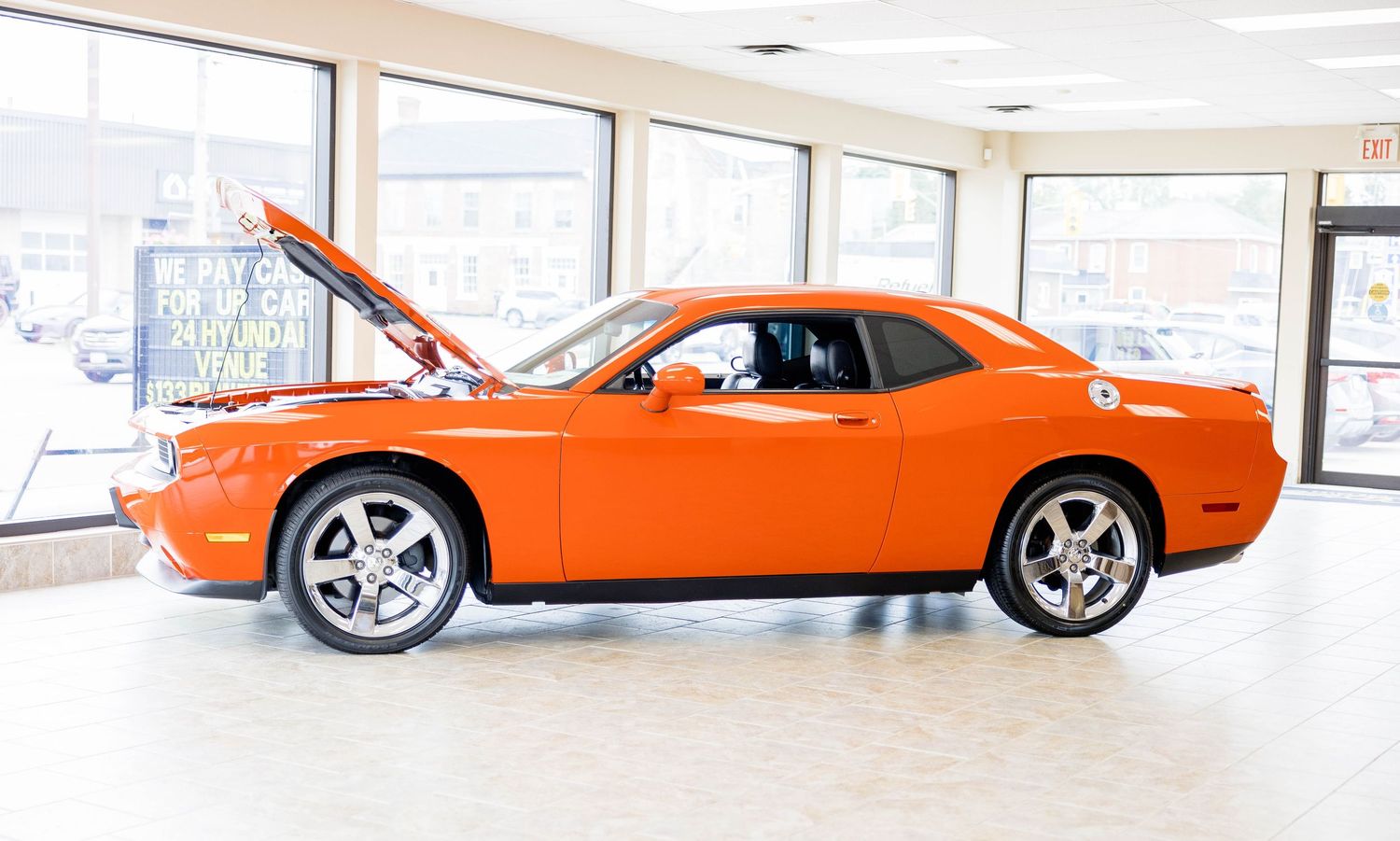 Orange Dodge Challenger with hood open displayed in a bright dealership showroom interior.