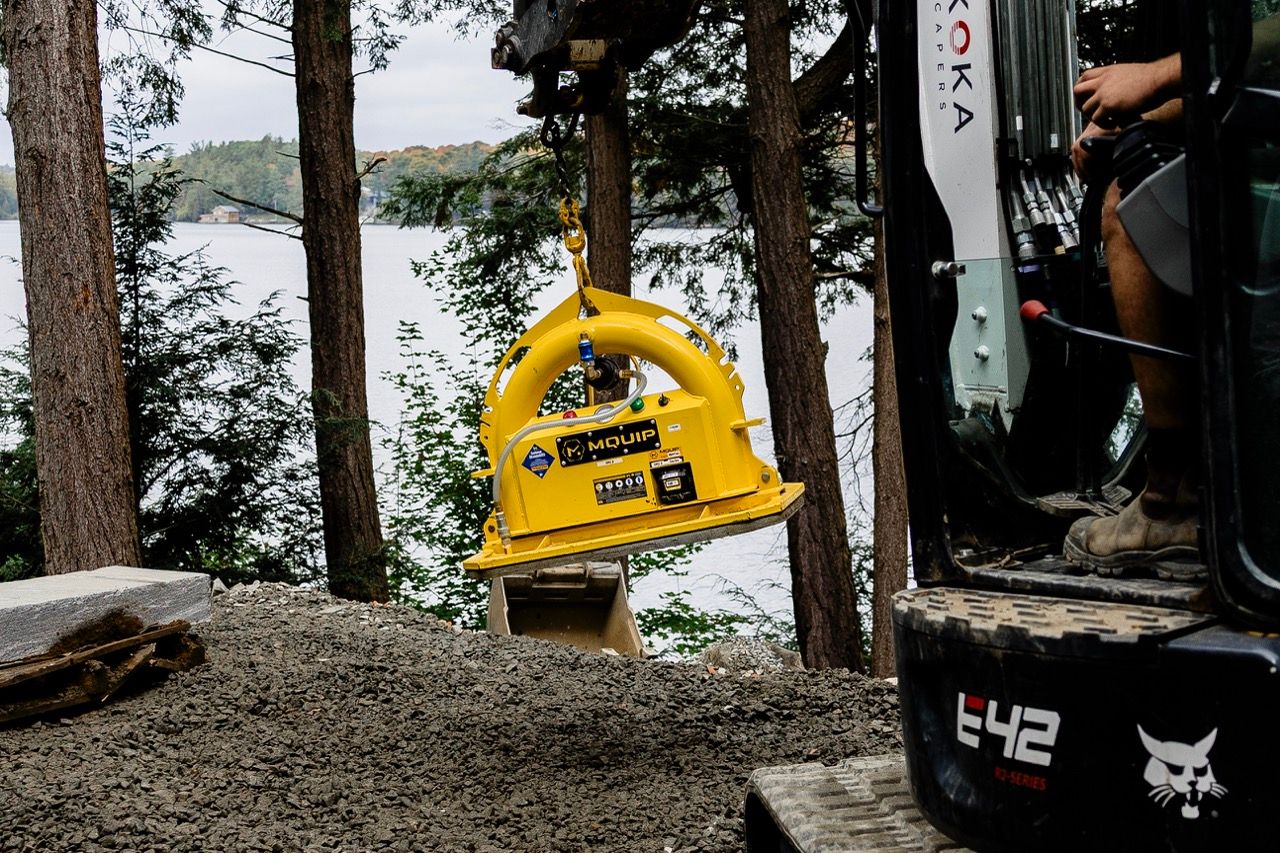 Yellow MQUIP vacuum lifter attachment suspended from Bobcat E42 excavator arm at forested lakeside worksite.