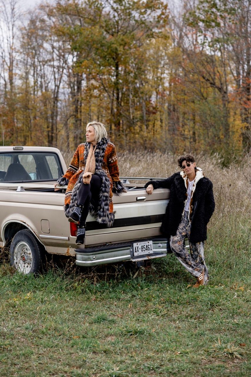 Two women in bohemian outfits pose by a vintage beige pickup truck in an autumn field.