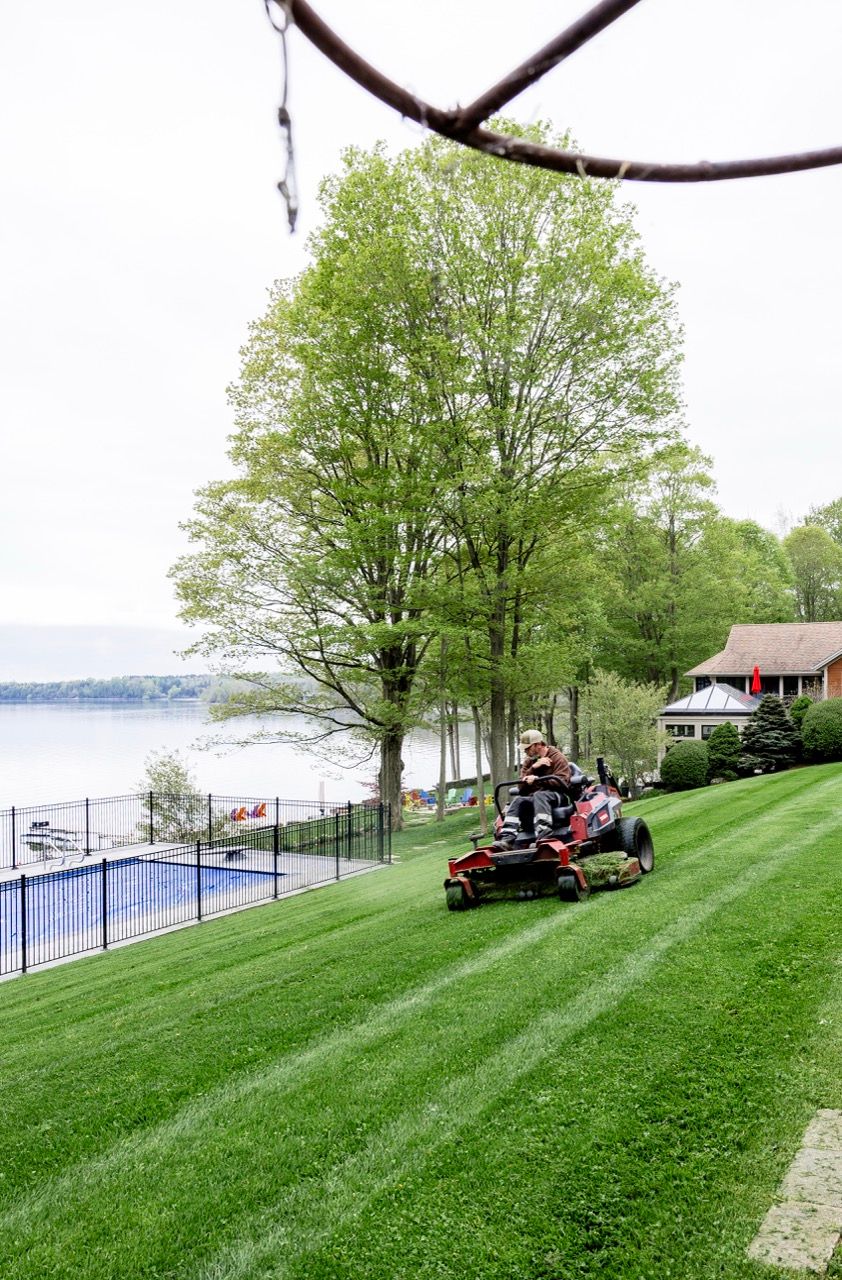 Landscaper on red zero-turn mower cutting striped green lawn near lakeside pool and trees.