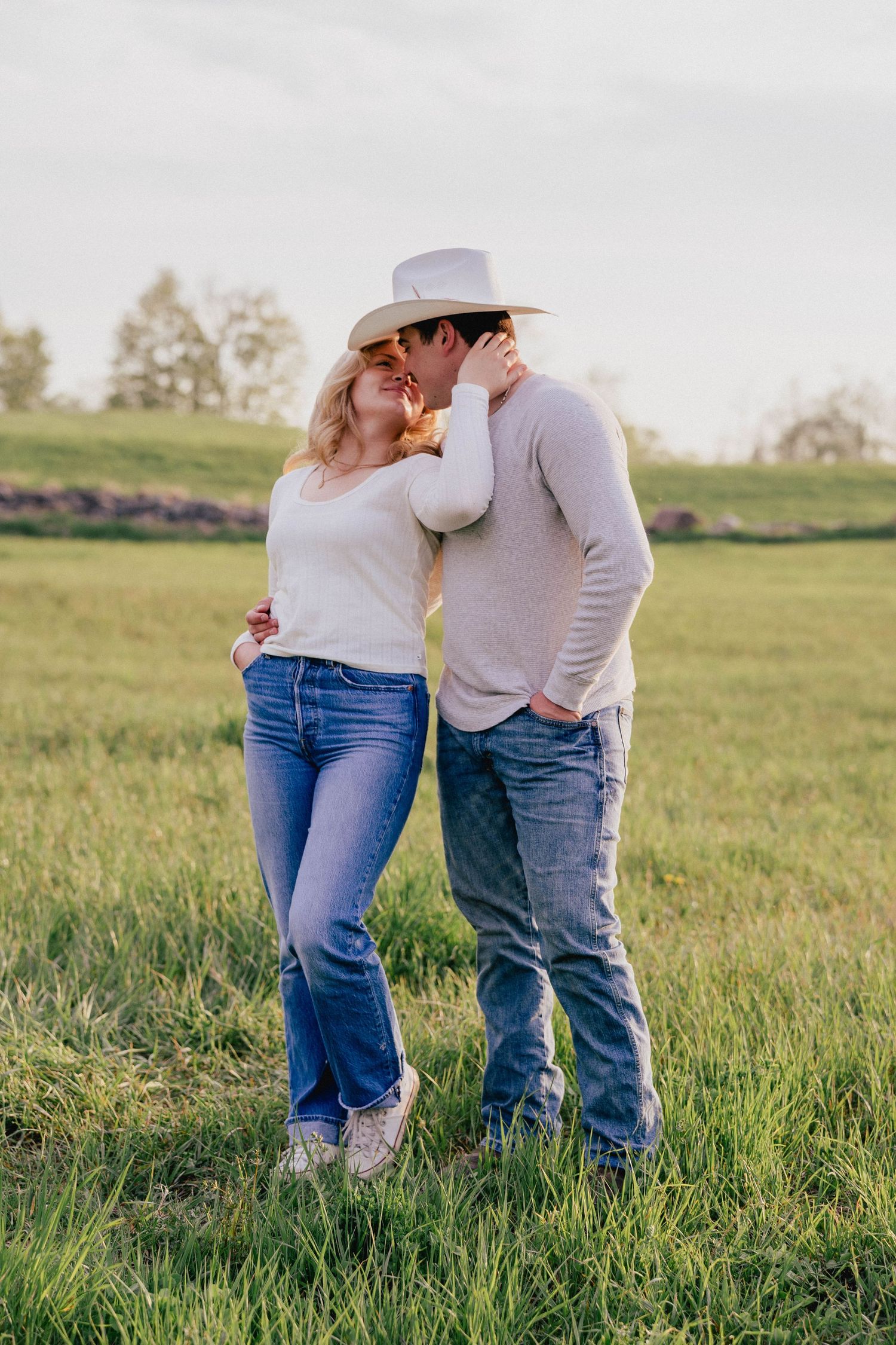 Couple sharing a romantic kiss in a sunlit green pasture, man wearing a white cowboy hat.