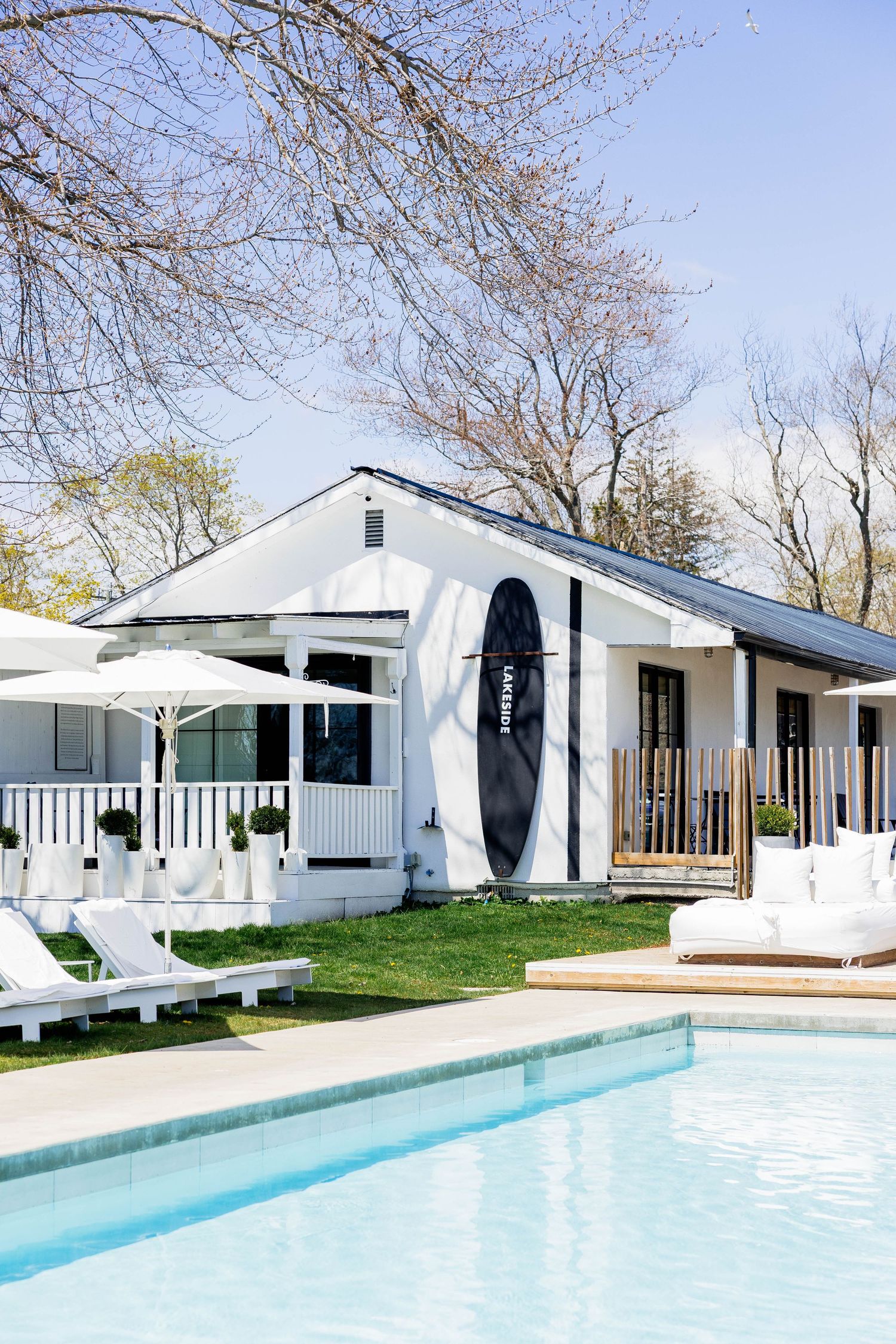 White pool house with surfboard, lounge chairs, and outdoor seating beside a sparkling swimming pool.