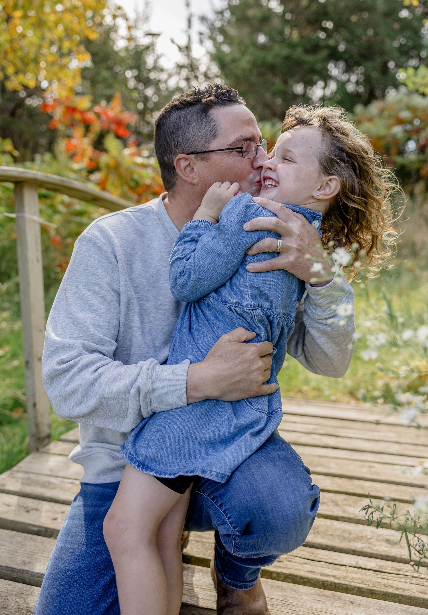 A man kisses a laughing curly-haired child on a wooden bridge surrounded by autumn foliage.
