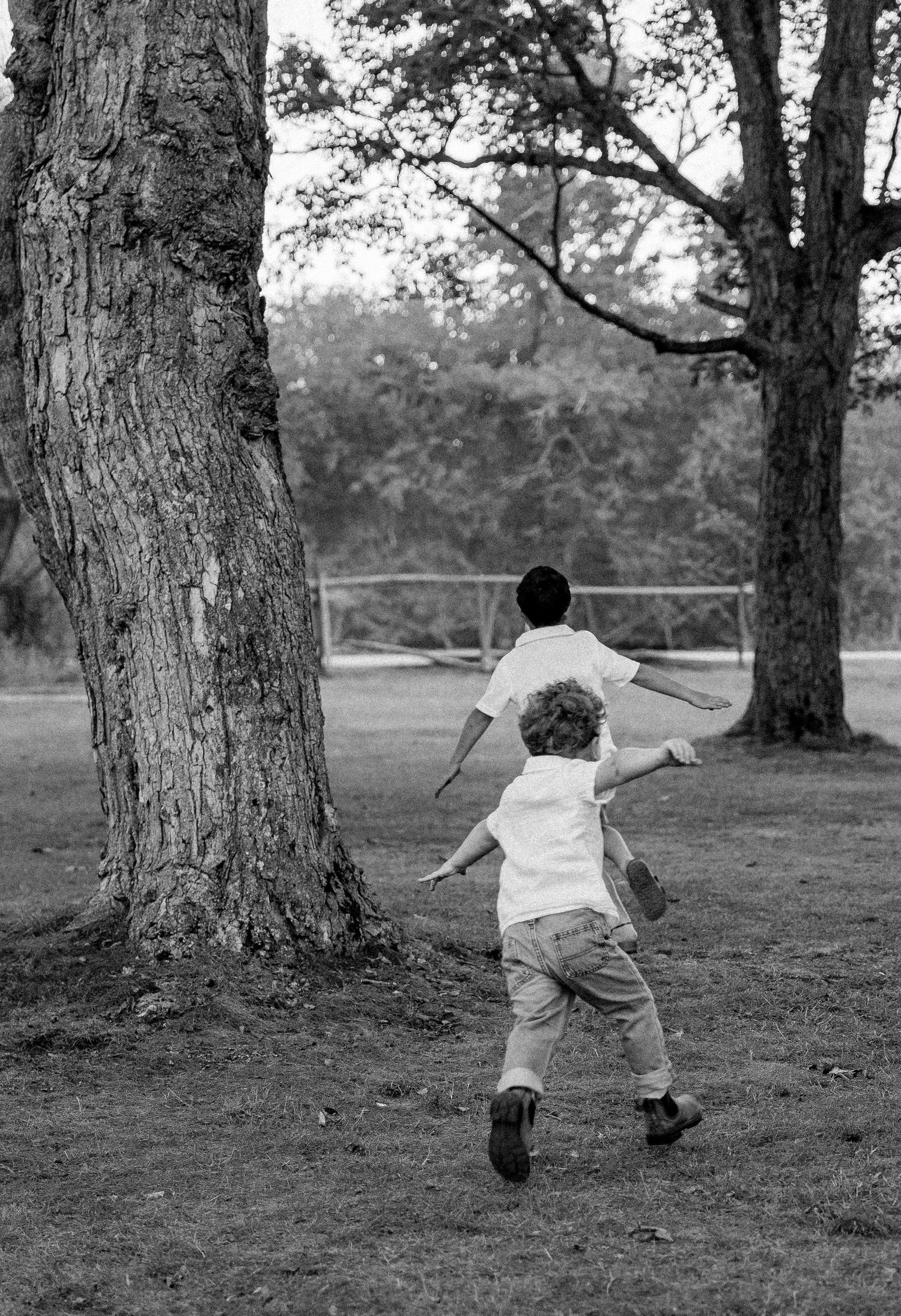Two children running between trees in a park, captured in black and white photography.