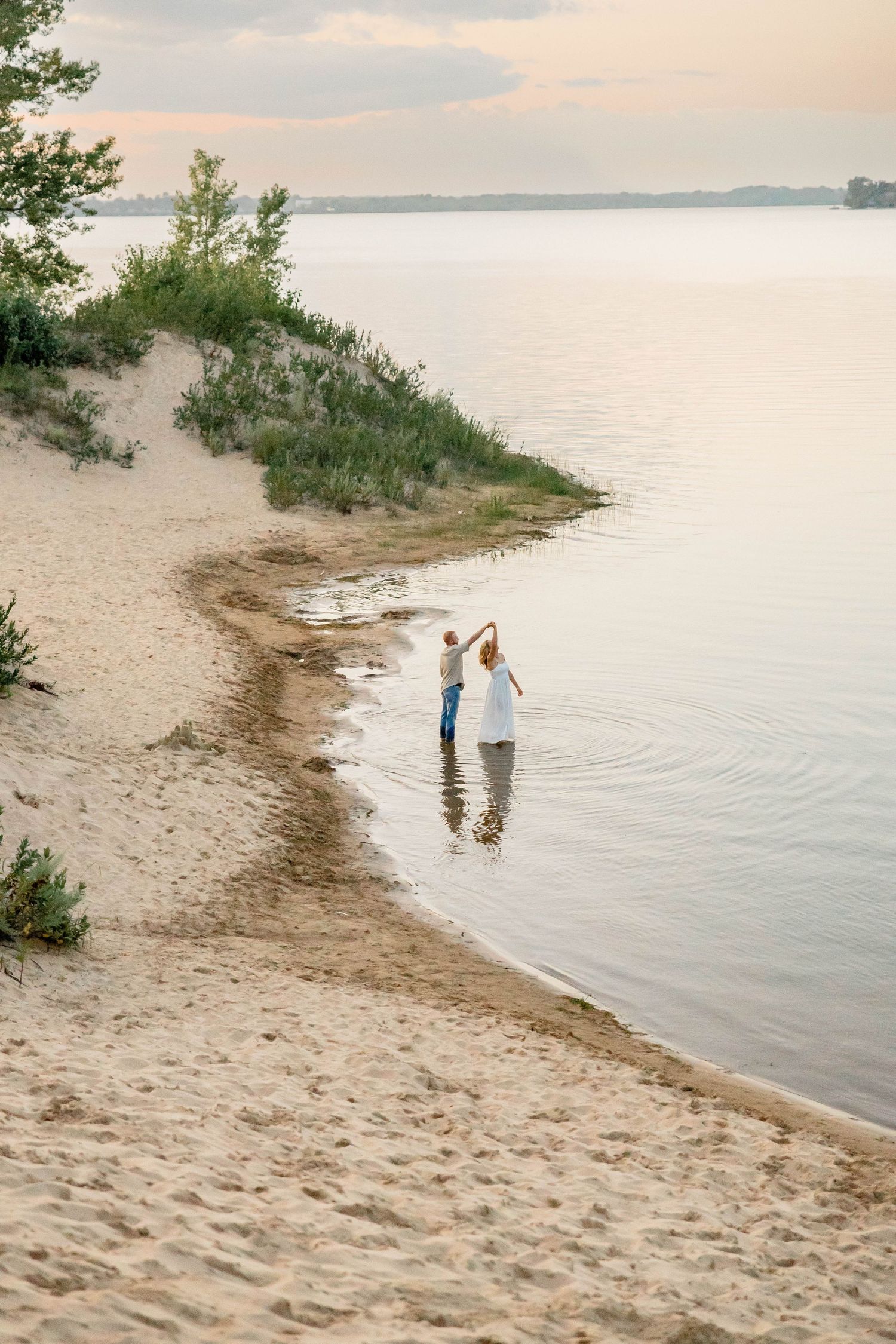 Couple in white attire wading in calm shallow water along a sandy beach shore at sunset.