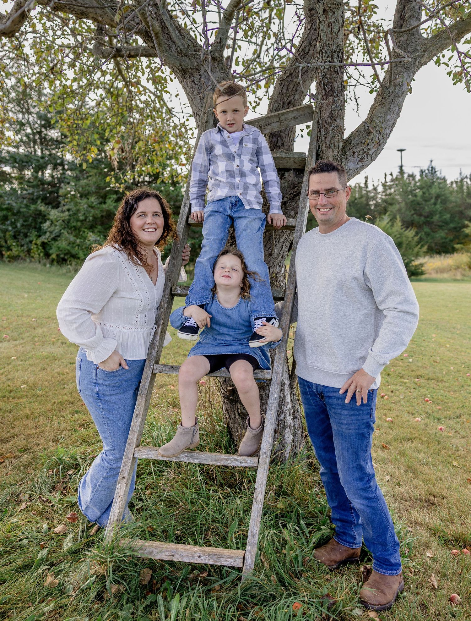 A smiling family of four poses with a rustic ladder against a large tree in a green outdoor setting.
