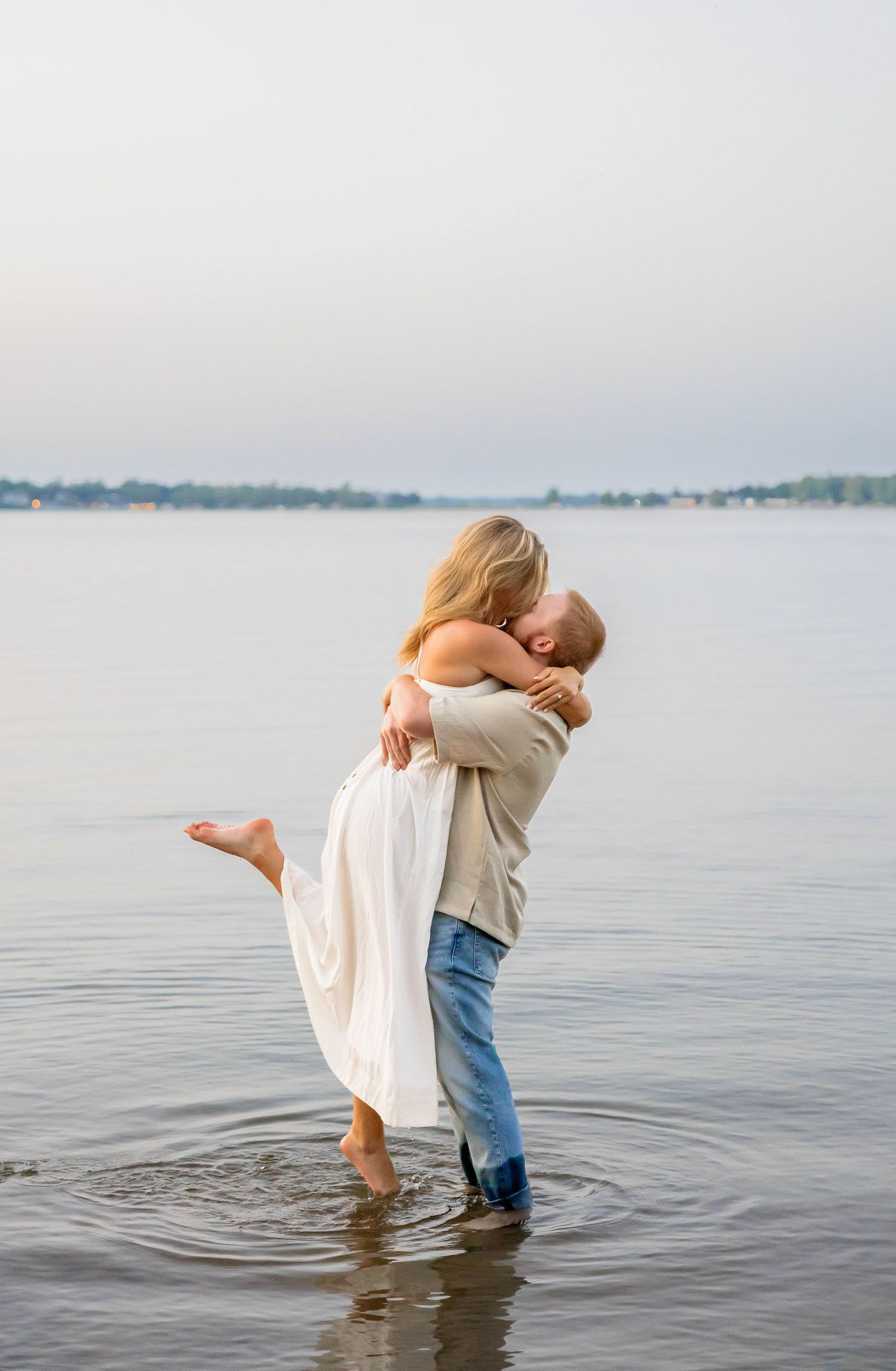 A couple shares a romantic kiss while standing in shallow water, the man lifting the woman in a white dress.