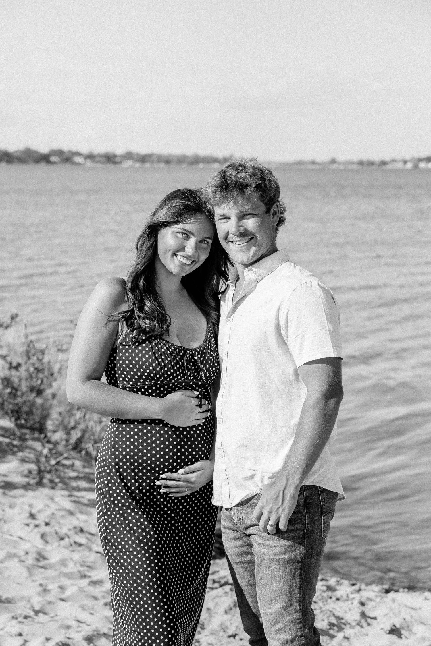 Black and white maternity photo of smiling couple on beach, woman in polka dot dress cradling baby bump, water in background.