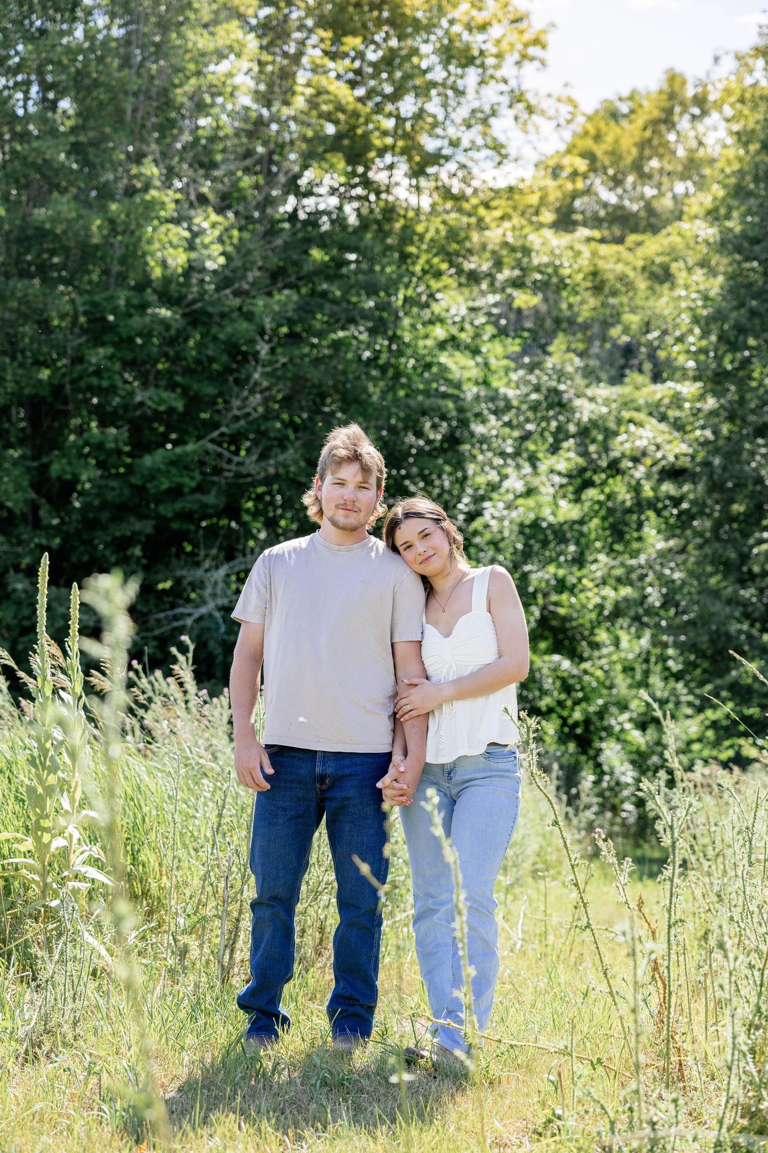 A couple stands together in a sunny green meadow, the woman resting her head on the man's shoulder.
