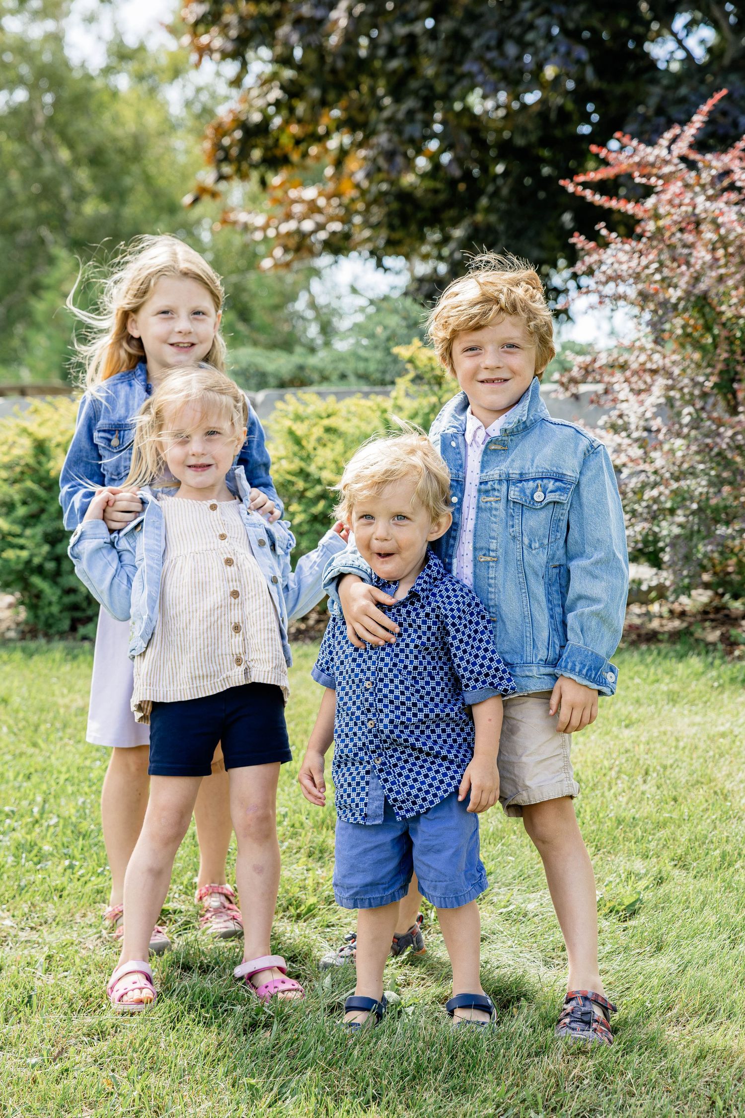 Four young children smiling together outdoors in a garden, wearing denim jackets on a sunny day.