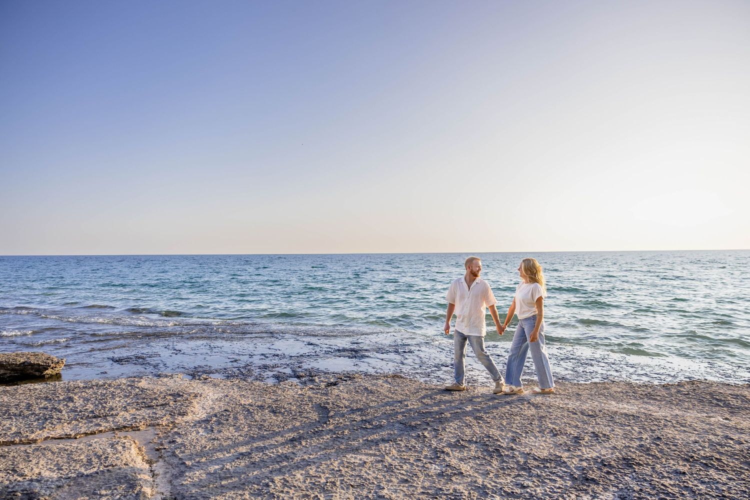 Elderly couple holding hands and walking along a rocky shoreline with calm blue ocean and clear sky.