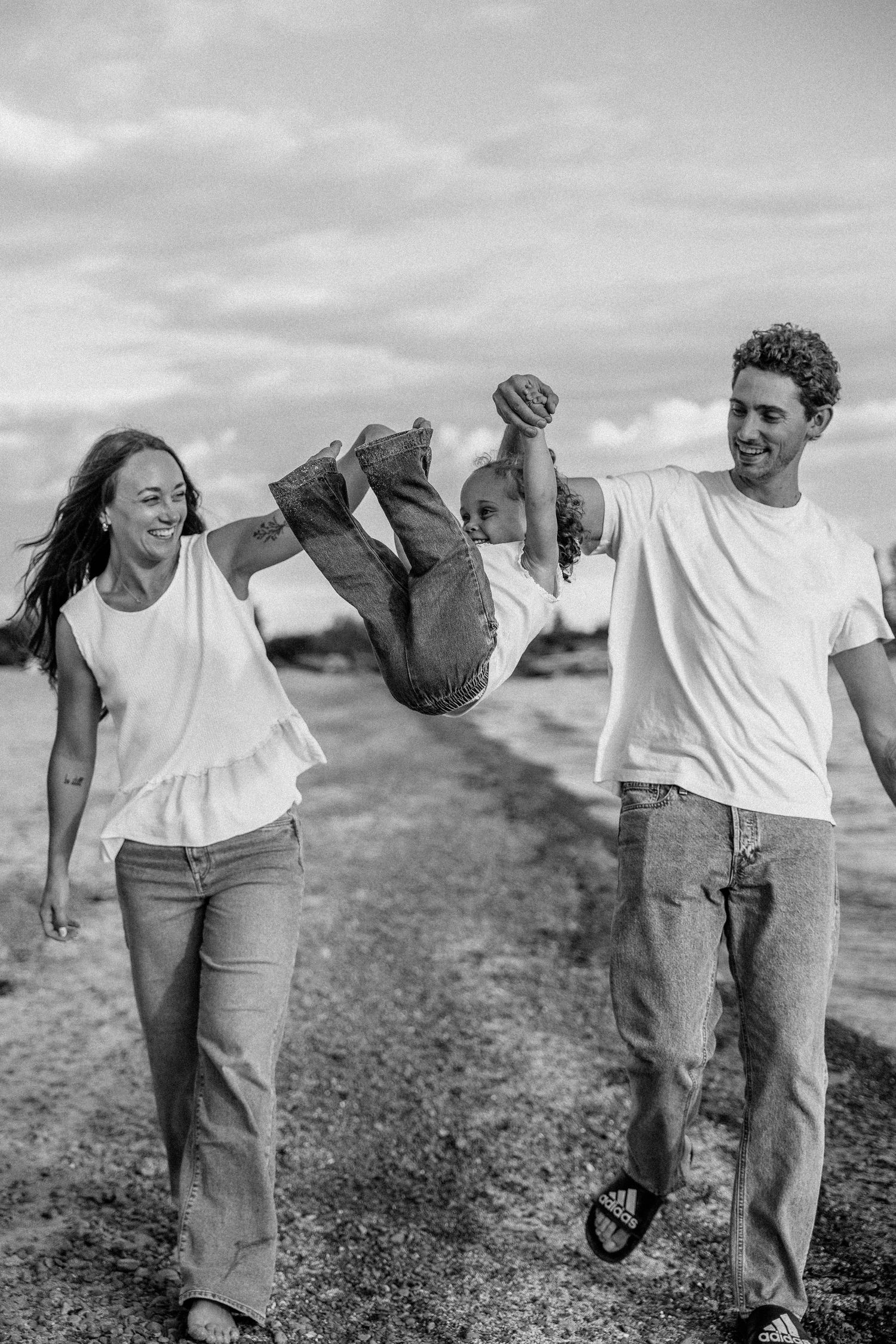 Black and white photo of happy family with parents swinging child between them while walking outdoors.