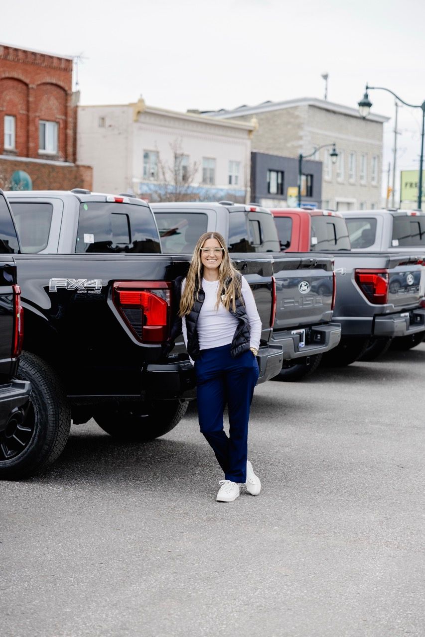 Smiling woman in navy pants and black vest standing beside black Ford F-150 trucks in a dealership lot.