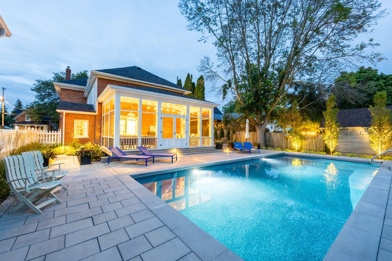 Backyard pool with illuminated pool house, patio furniture, and lush landscaping at twilight.