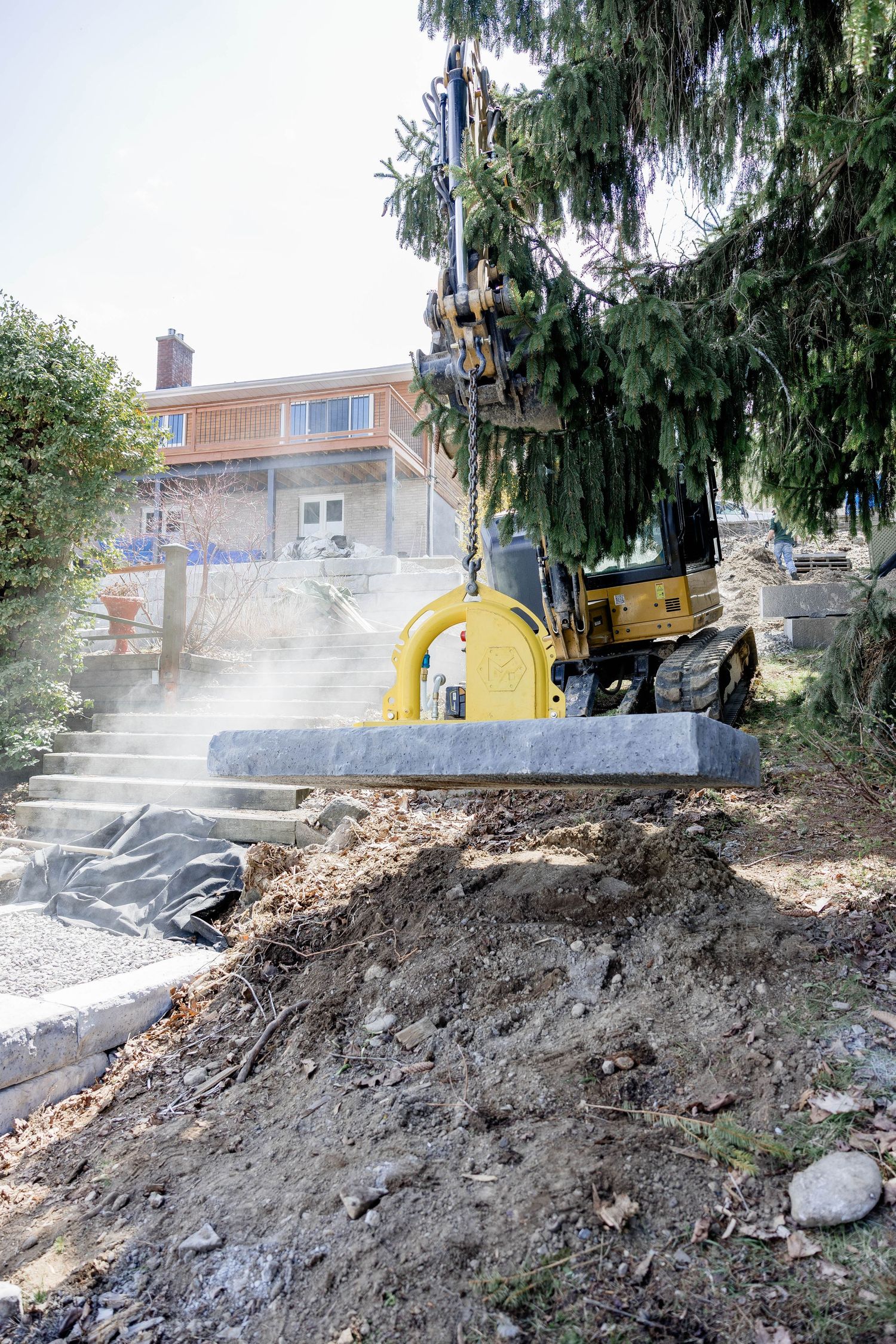 Yellow excavator lifting a large stone slab in a residential garden landscape.