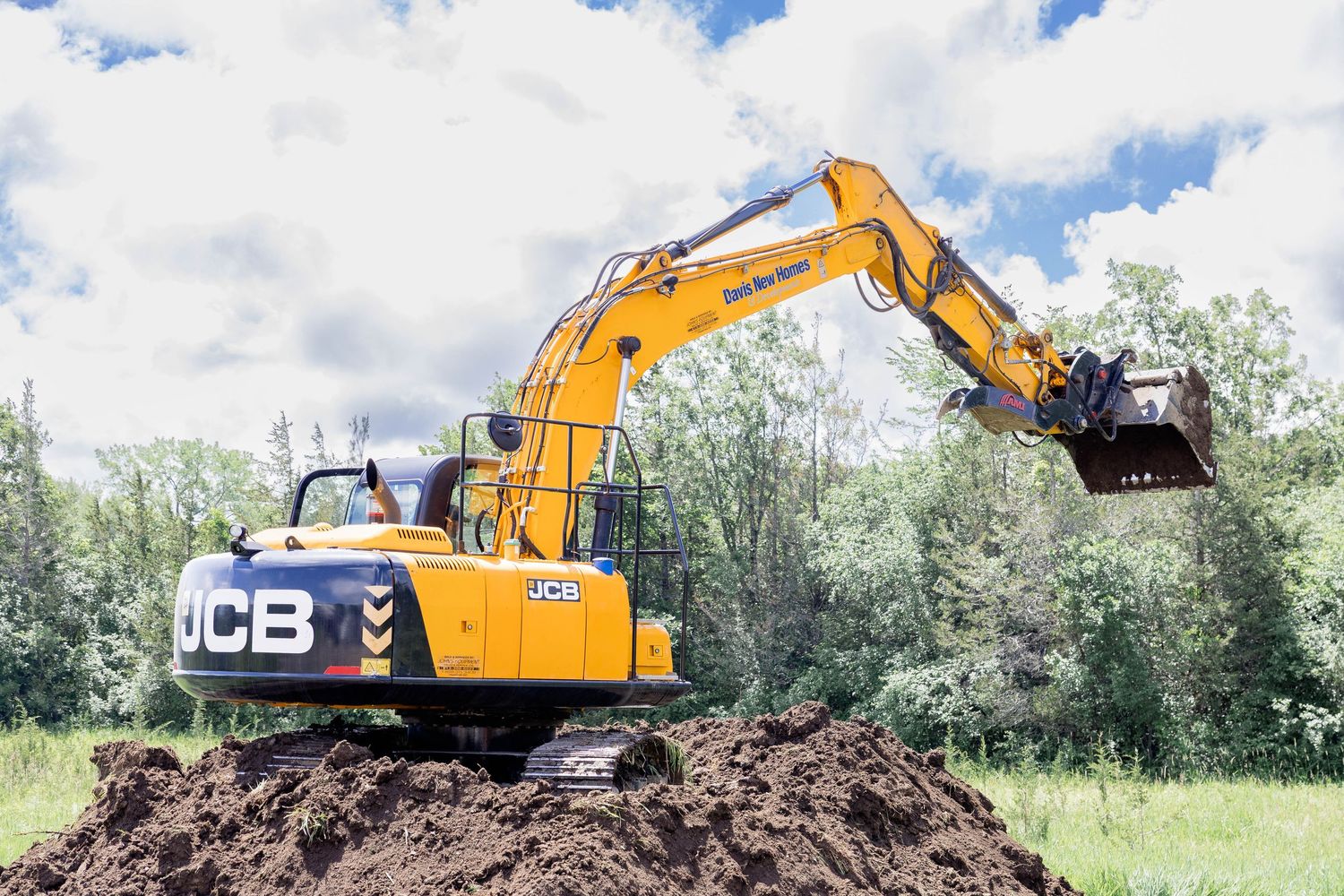 JCB yellow excavator moves earth on a construction site surrounded by lush green trees.