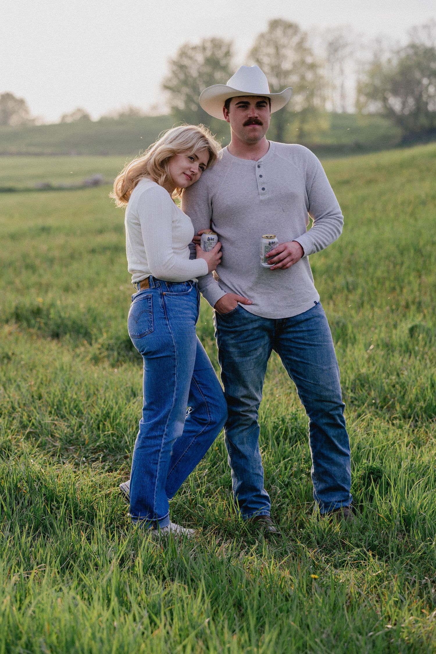 Couple posing together in a green field at sunset, man wearing cowboy hat holding a can.