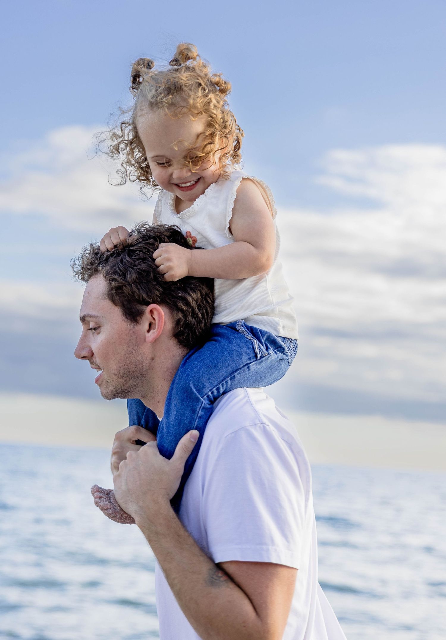 A happy young girl sits on a man's shoulders at the beach, smiling joyfully under a blue sky.