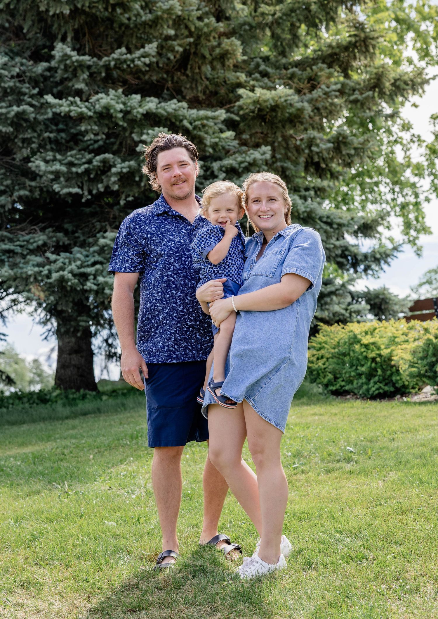 Happy family of three posing outdoors on green grass with evergreen trees, wearing coordinating blue outfits.