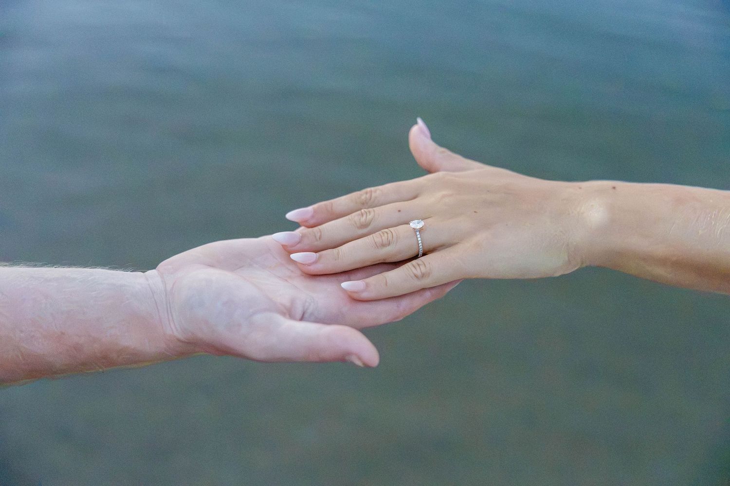 Two hands reaching toward each other over water, woman wearing a diamond engagement ring on her finger.