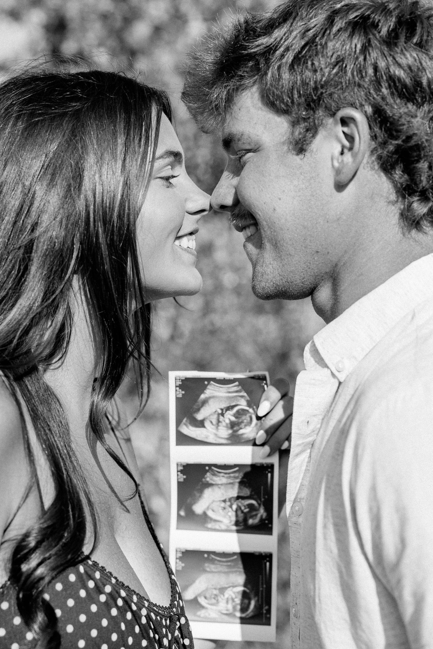 Black and white photo of a smiling couple touching noses while holding ultrasound photos, announcing their pregnancy.