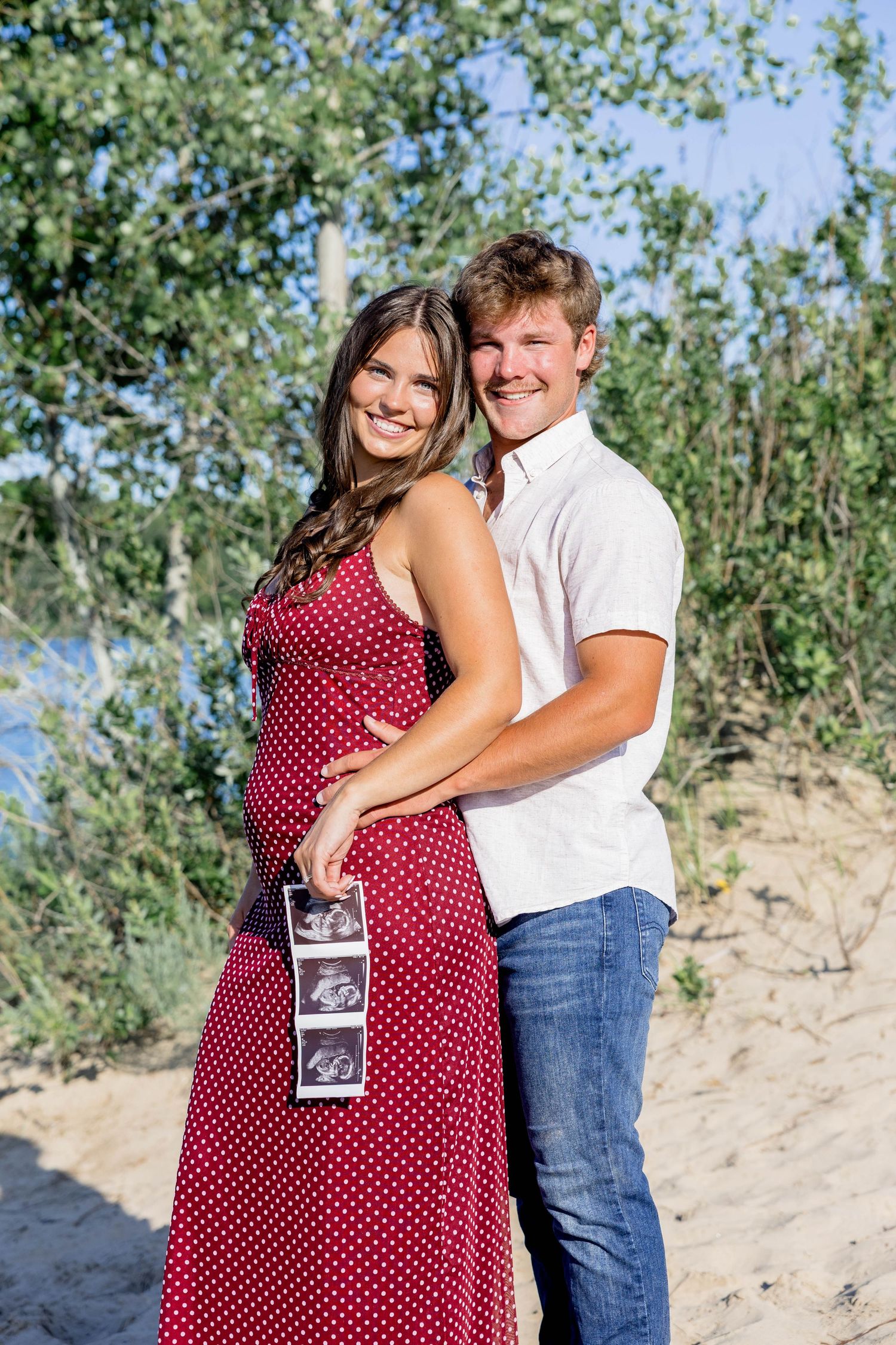 Happy couple holding ultrasound photos outdoors, pregnant woman in red polka dot dress embraced by smiling man in white shirt.