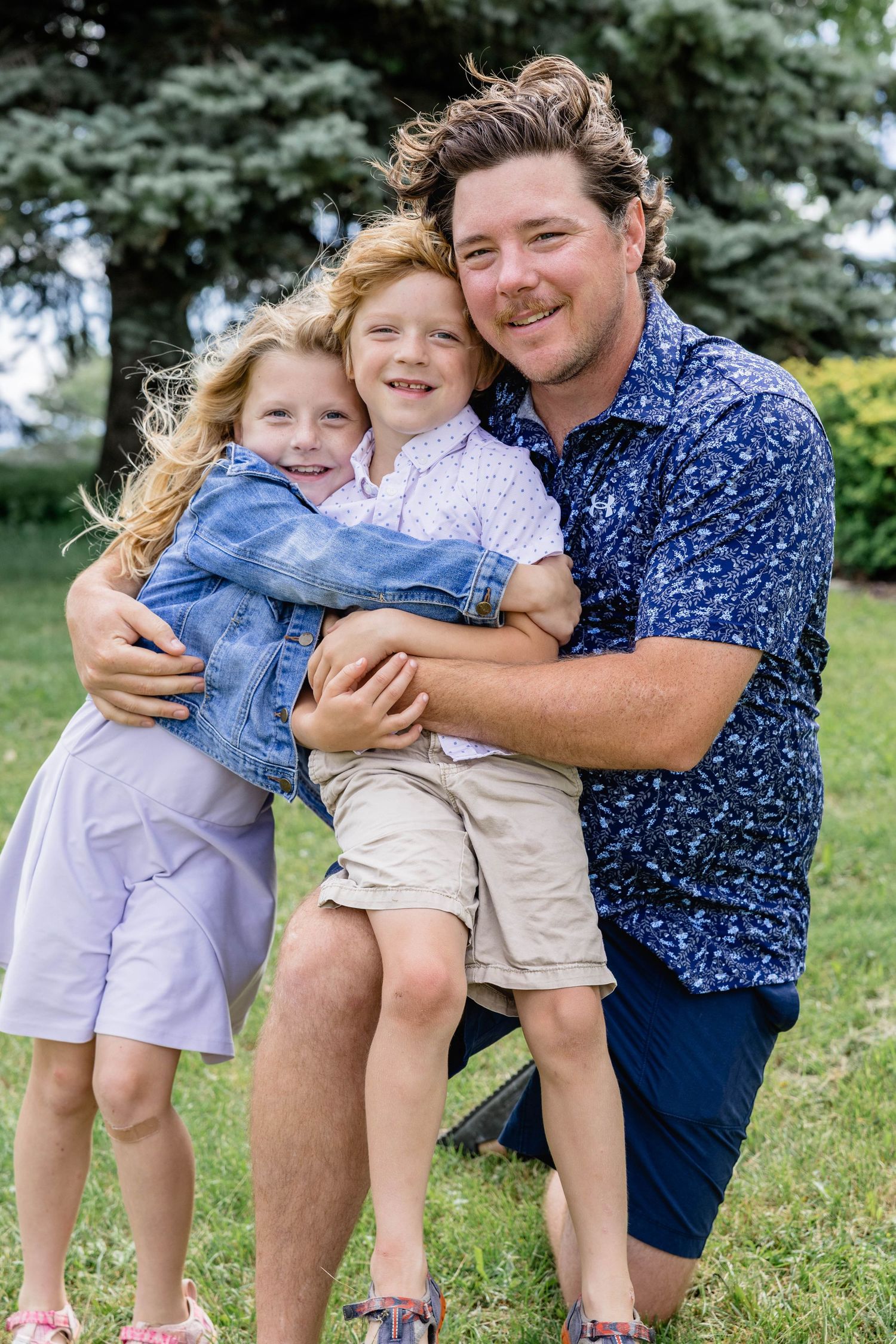 A smiling man kneels outdoors hugging two young redheaded children on a sunny day in a green park.