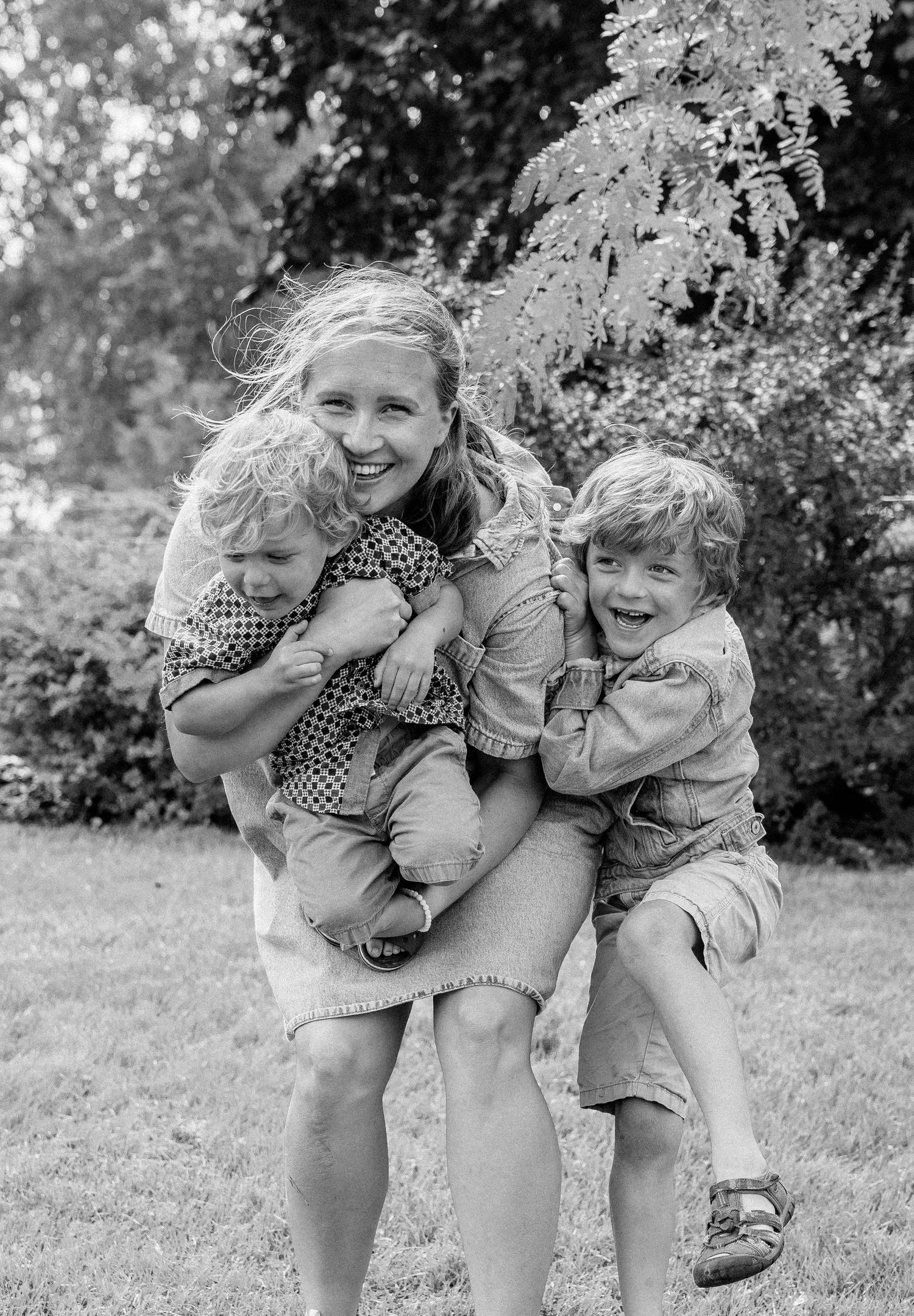 Black and white photo of a smiling mother carrying two young blonde boys in a lush garden setting.