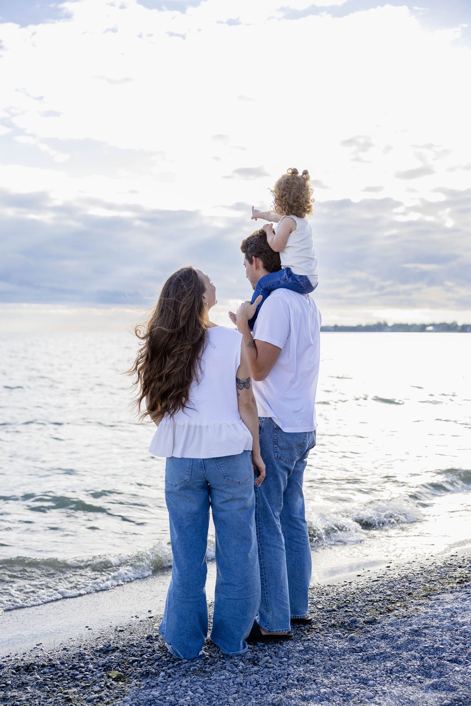 Family of three standing on rocky beach shoreline, parents in white tops and jeans with toddler on fathers shoulders.