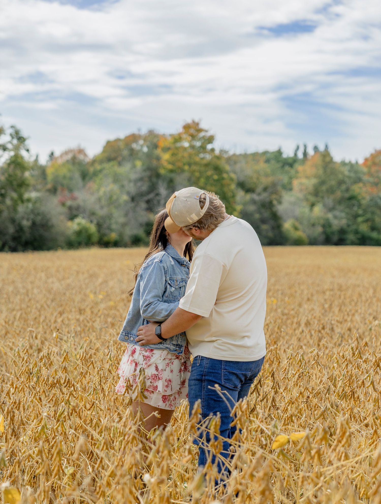 Couple kissing in golden wheat field with autumn trees and cloudy sky in romantic outdoor engagement photo.