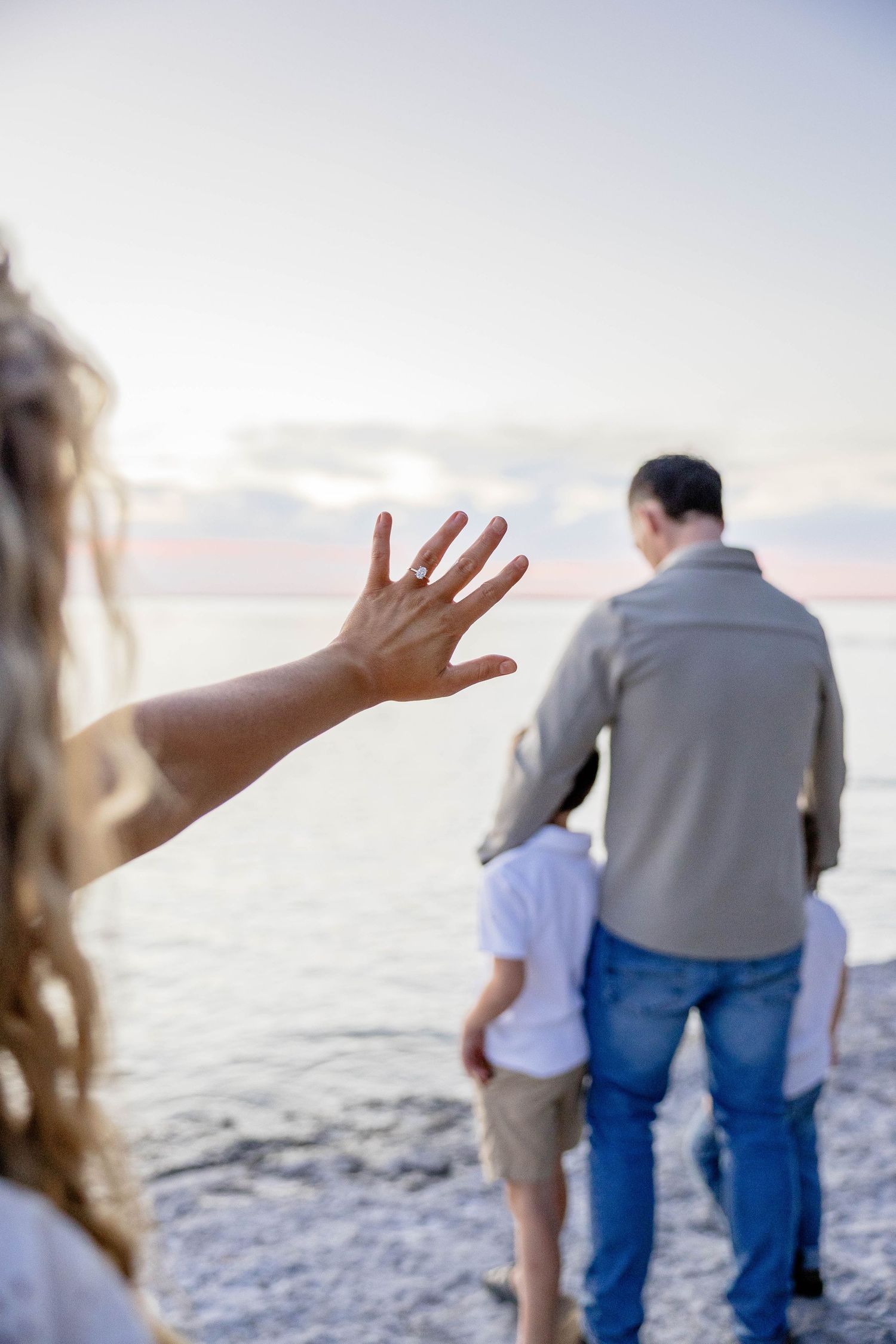 Woman waving hand on beach while man and child stand near ocean at sunset.