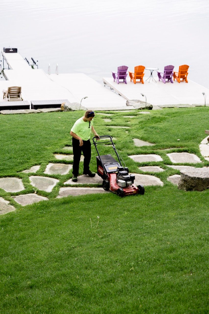 Landscaper mowing green lawn with red push mower near stone path and colorful lakeside chairs.
