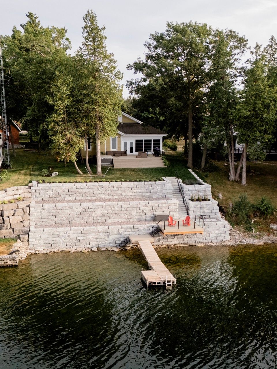 A lakeside cottage with a large stone retaining wall, wooden dock, and lush green trees at dusk.