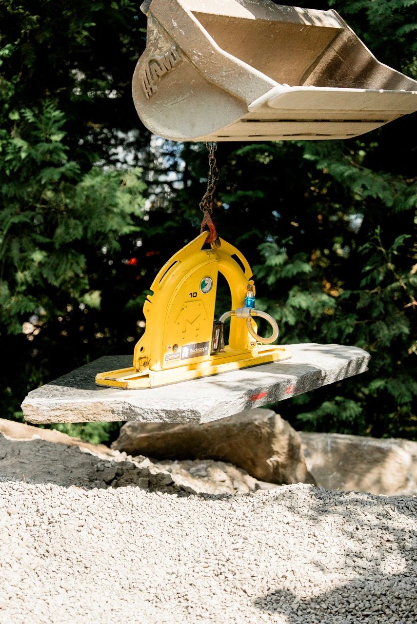Yellow excavator stone lifter clamp gripping granite slab over gravel at outdoor site.