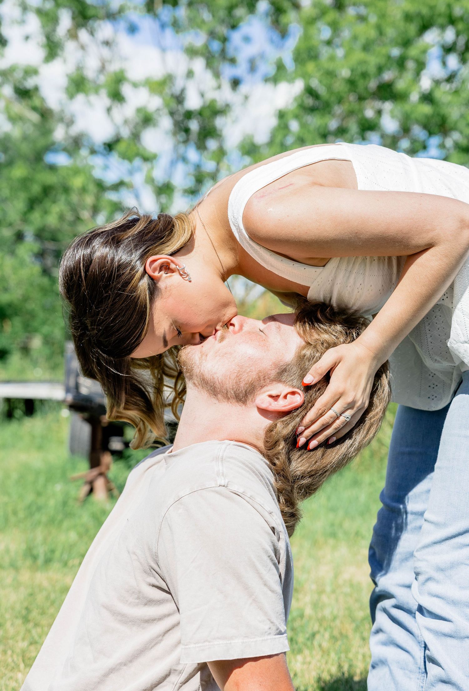 A couple shares a romantic kiss outdoors in a sunny green park, with the woman leaning down to kiss the man.