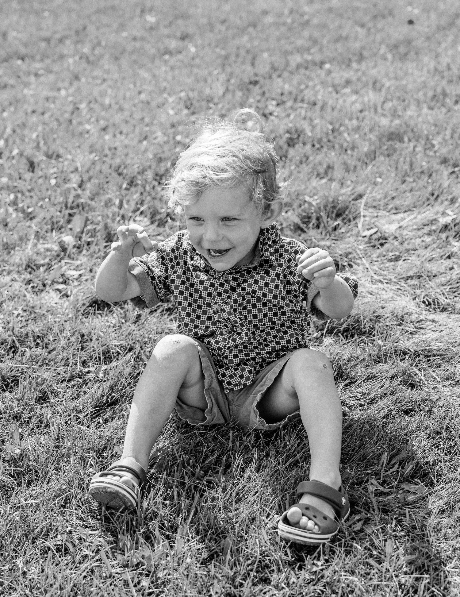 Happy young child sitting on grass, laughing with fists raised, wearing patterned outfit and sandals, black and white photo.