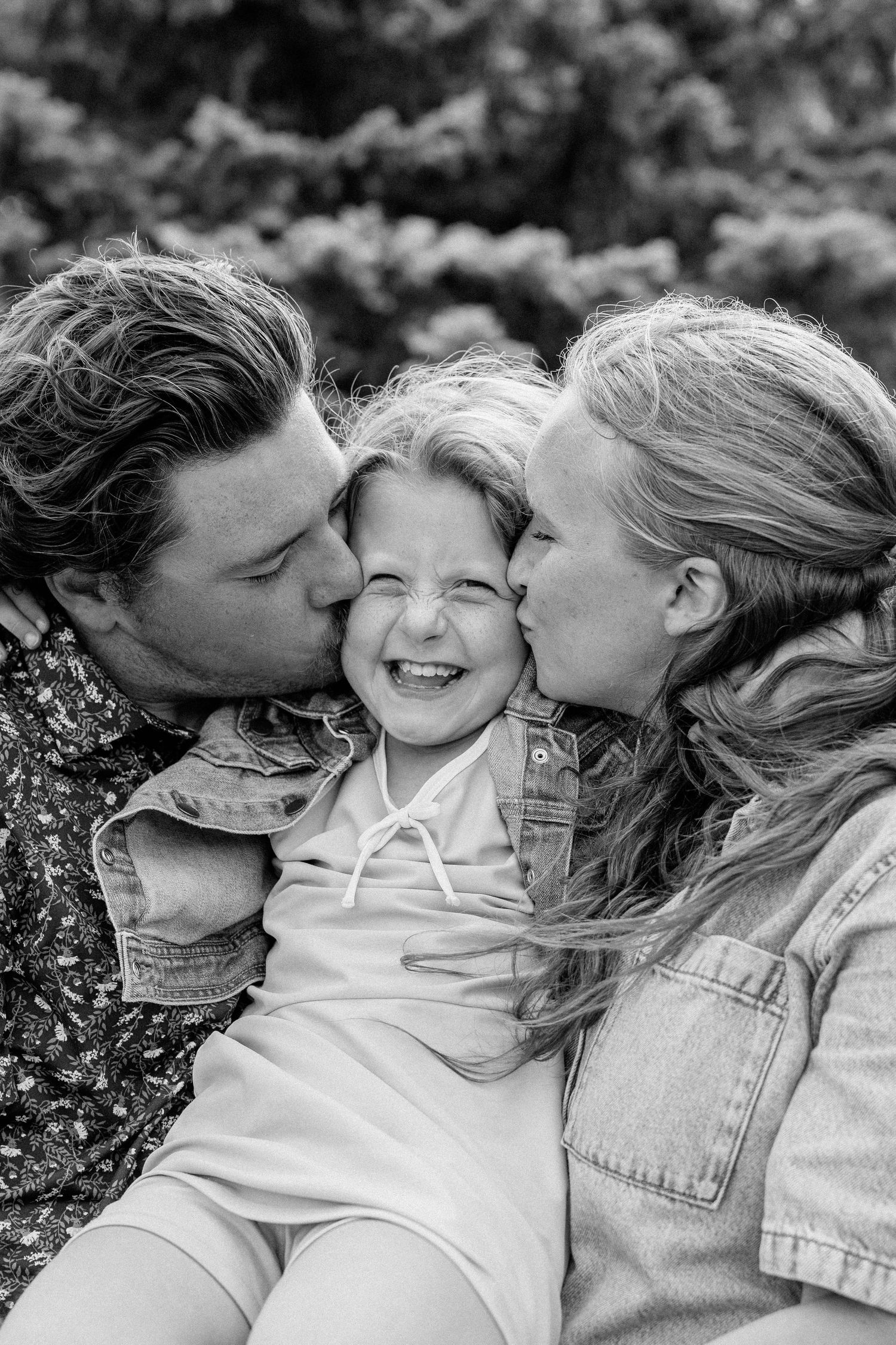Black and white photo of two parents kissing their laughing young child on the cheeks outdoors.