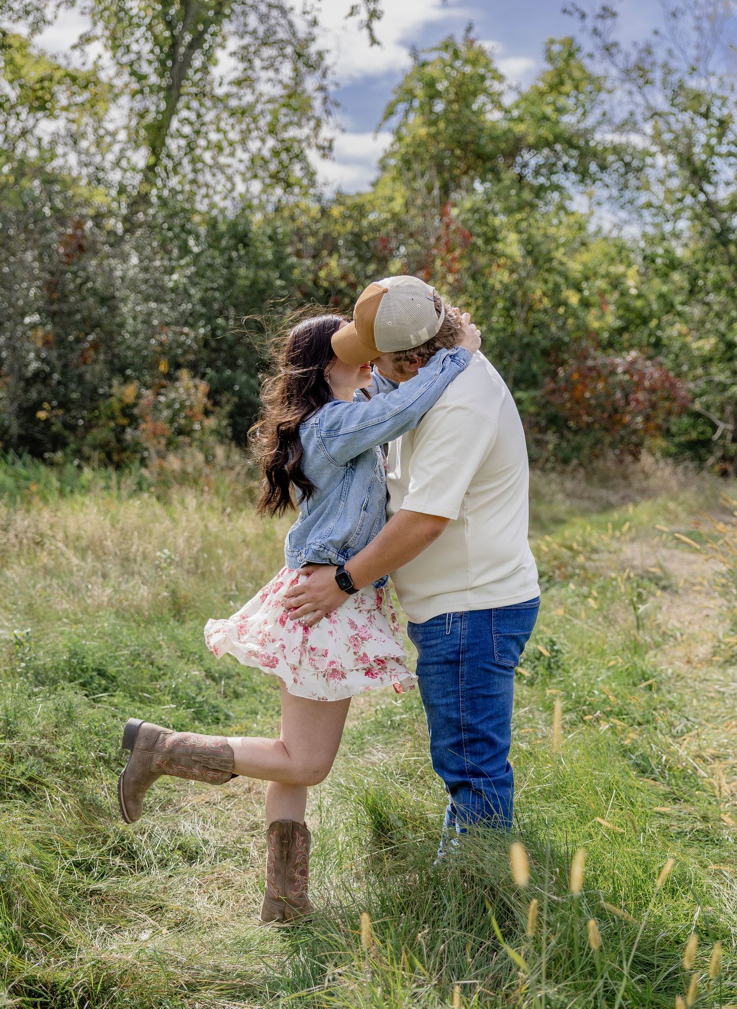 Couple sharing a romantic kiss in a green meadow, woman in floral skirt and denim jacket, man in cream shirt and jeans.