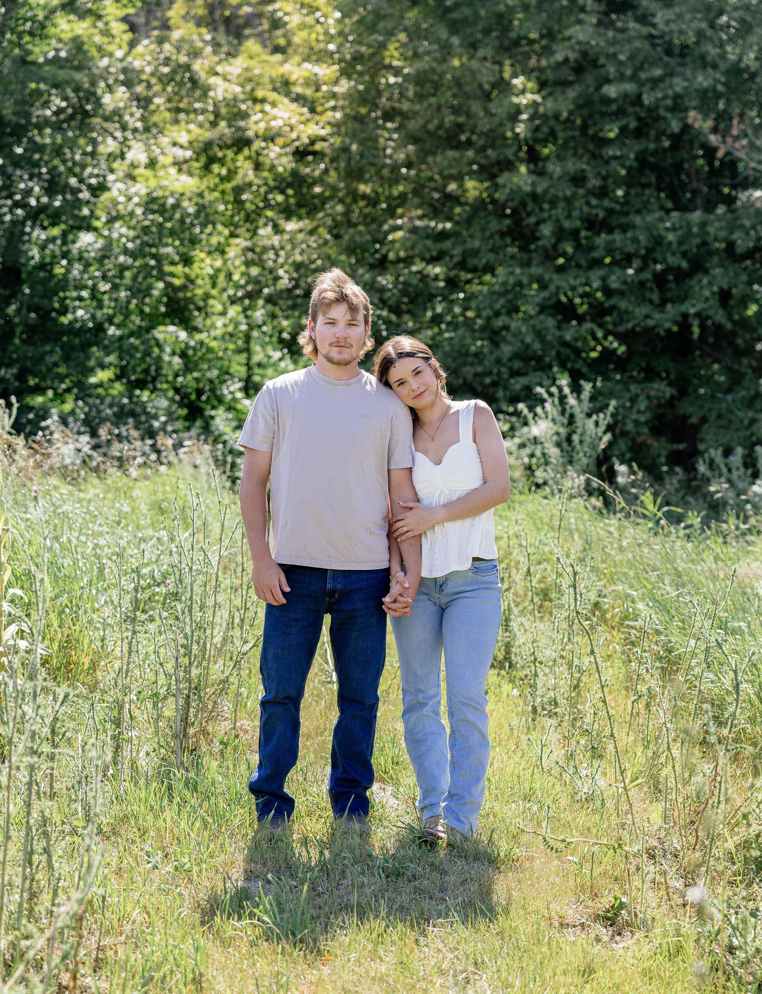 A couple holds hands and poses together in a lush green meadow surrounded by trees on a sunny day.