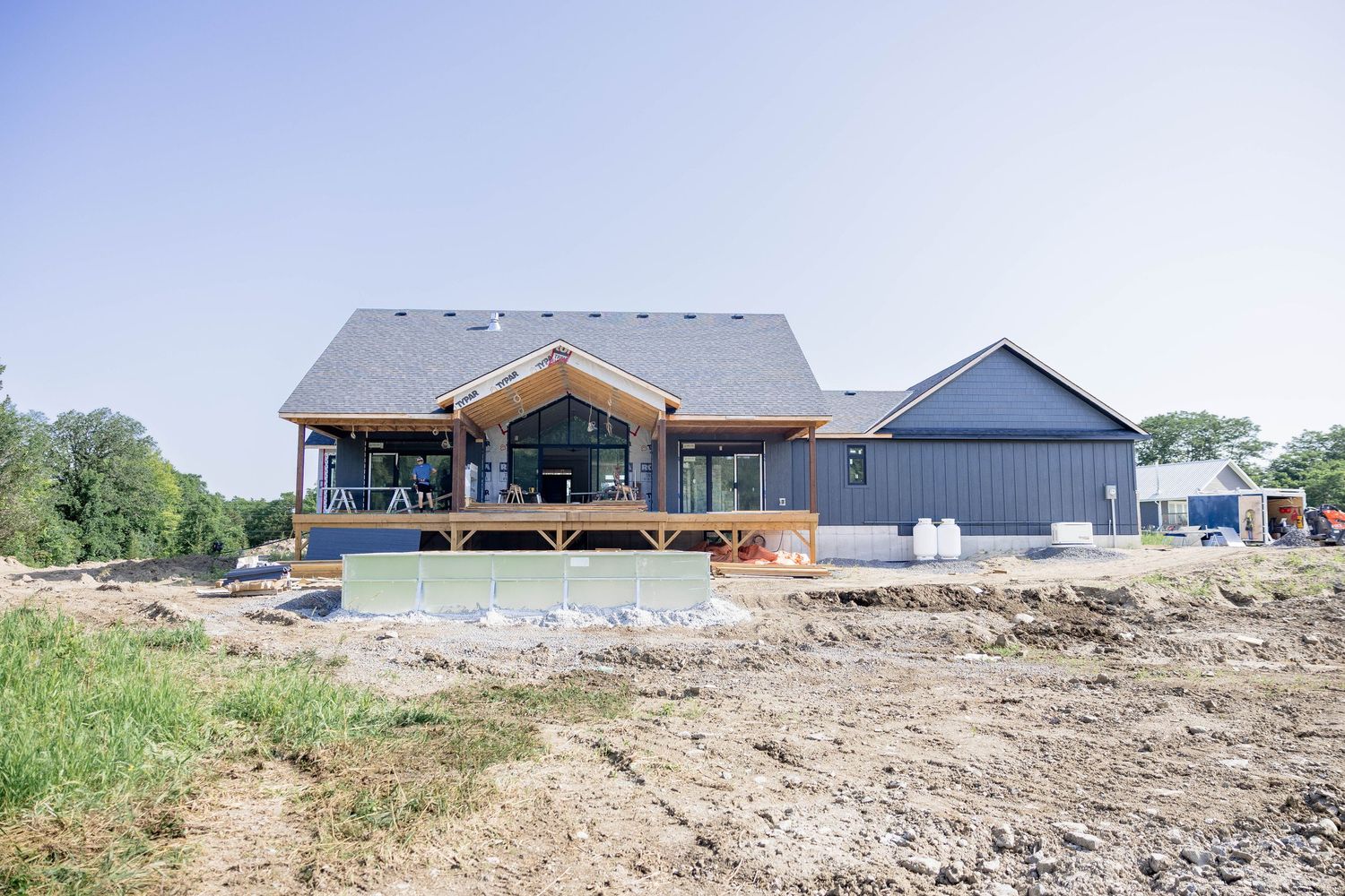 Modern farmhouse under construction featuring large windows and covered porch overlooking cleared land.