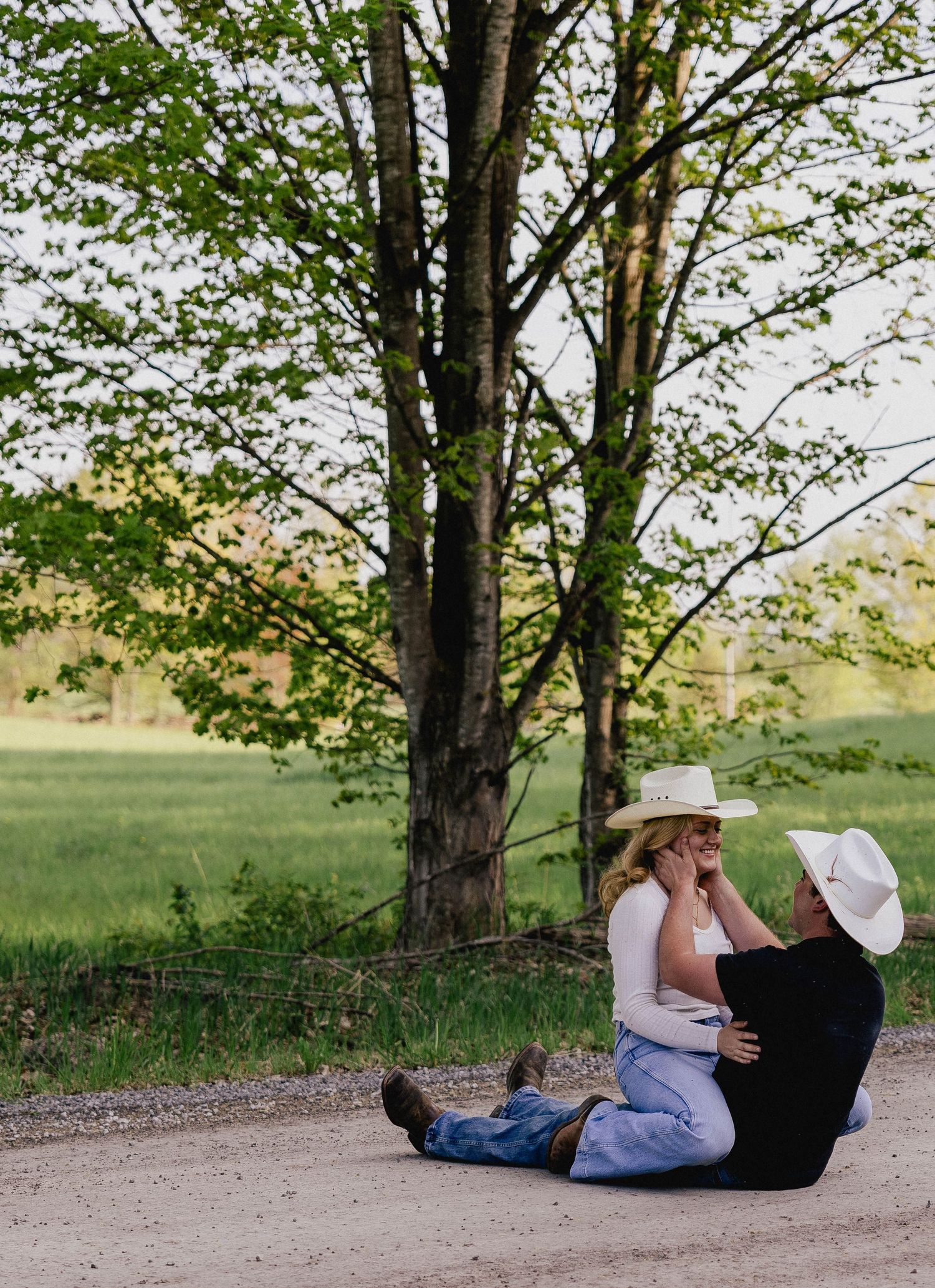 Couple sitting on dirt road laughing together, woman in cowboy hat, green trees in background.
