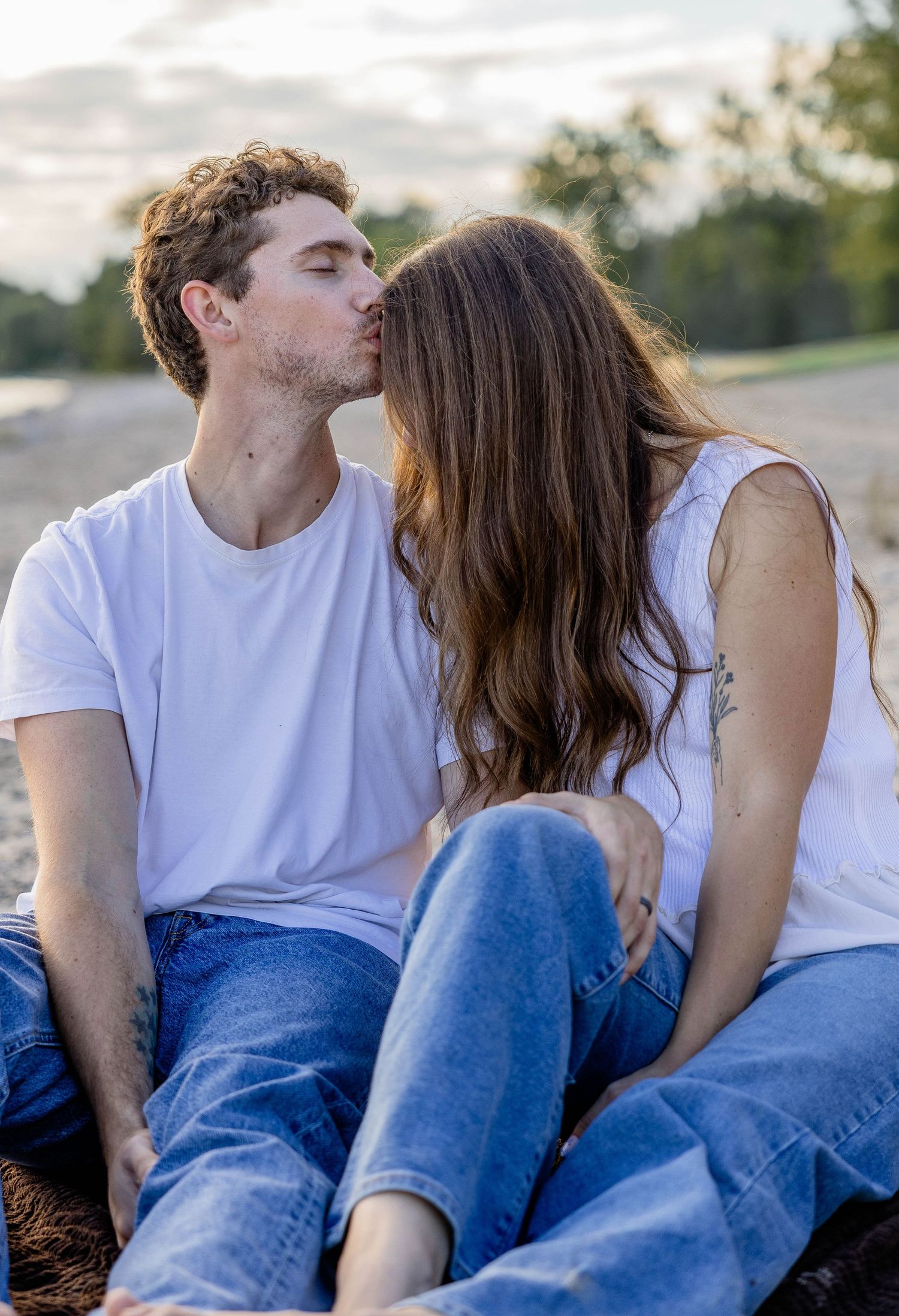 A young couple sharing a kiss while sitting outdoors, both dressed in white shirts and blue jeans at sunset.