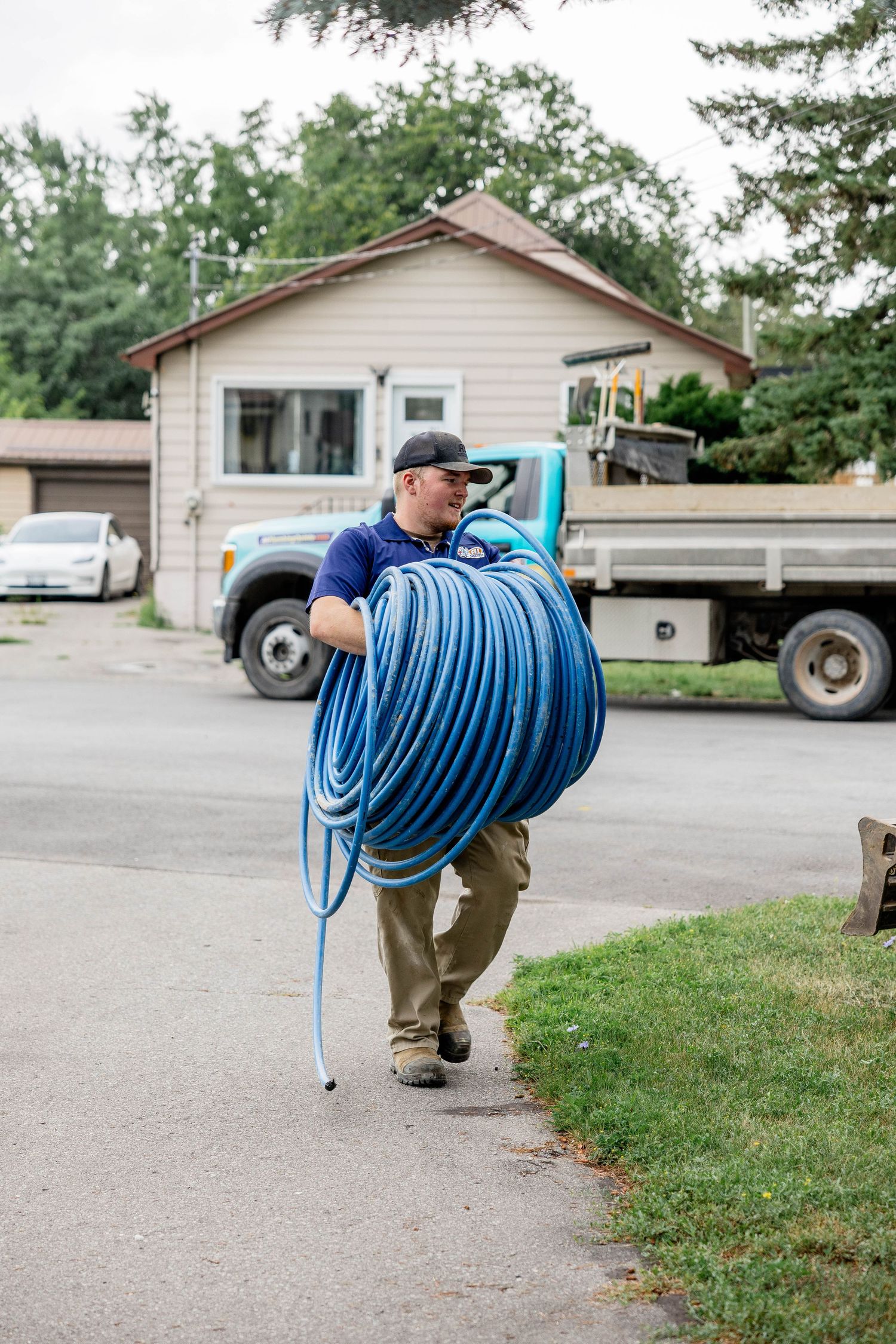 Worker carrying a large coiled blue hose while walking down a residential driveway near a flatbed truck.