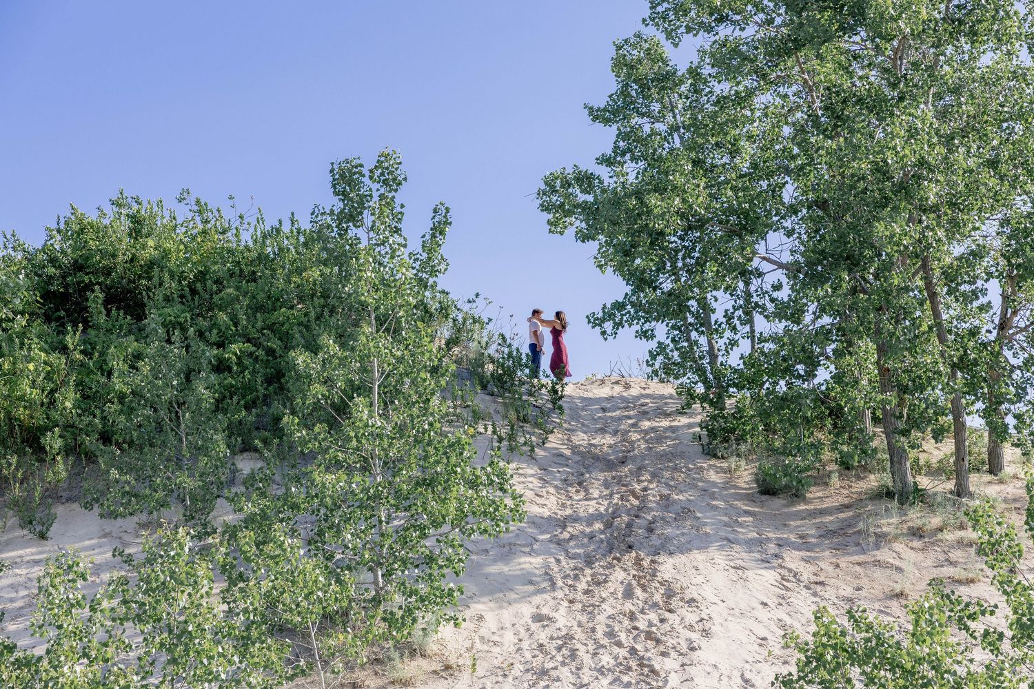 Two people stand atop a sandy dune trail surrounded by lush green trees under a clear blue sky.