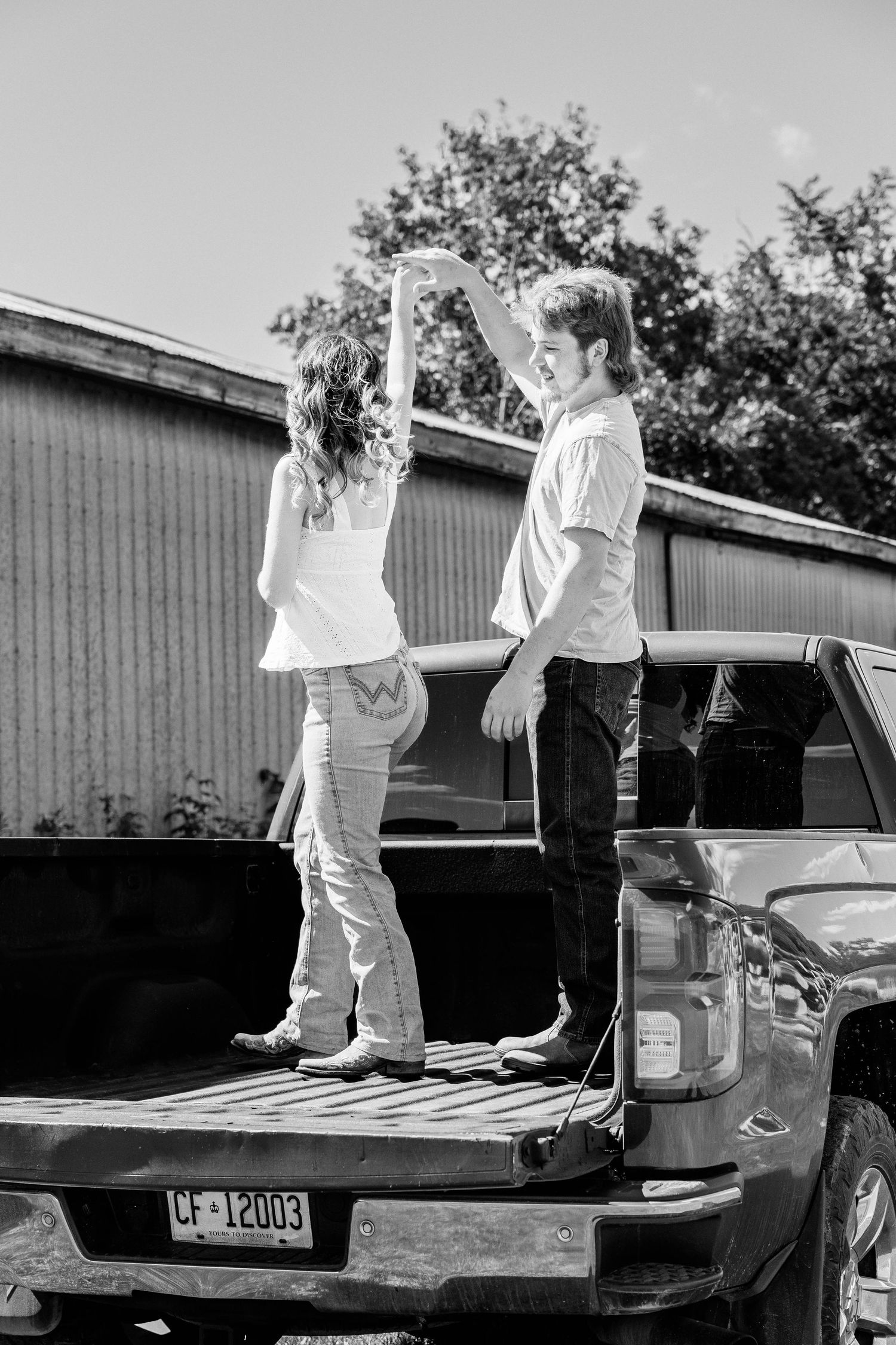 Black and white photo of a young couple dancing on a pickup truck bed outdoors near a barn.