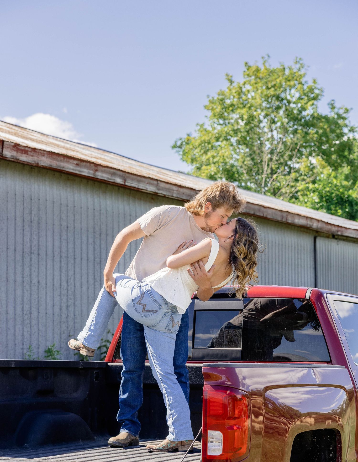 Couple sharing a romantic dip kiss beside a red pickup truck near a rustic barn on a sunny day.