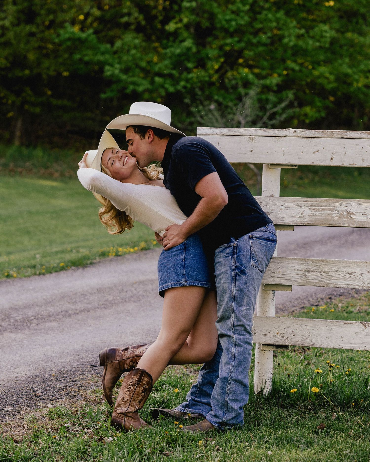 A couple shares a passionate dip kiss near a white wooden fence on a rural country road.
