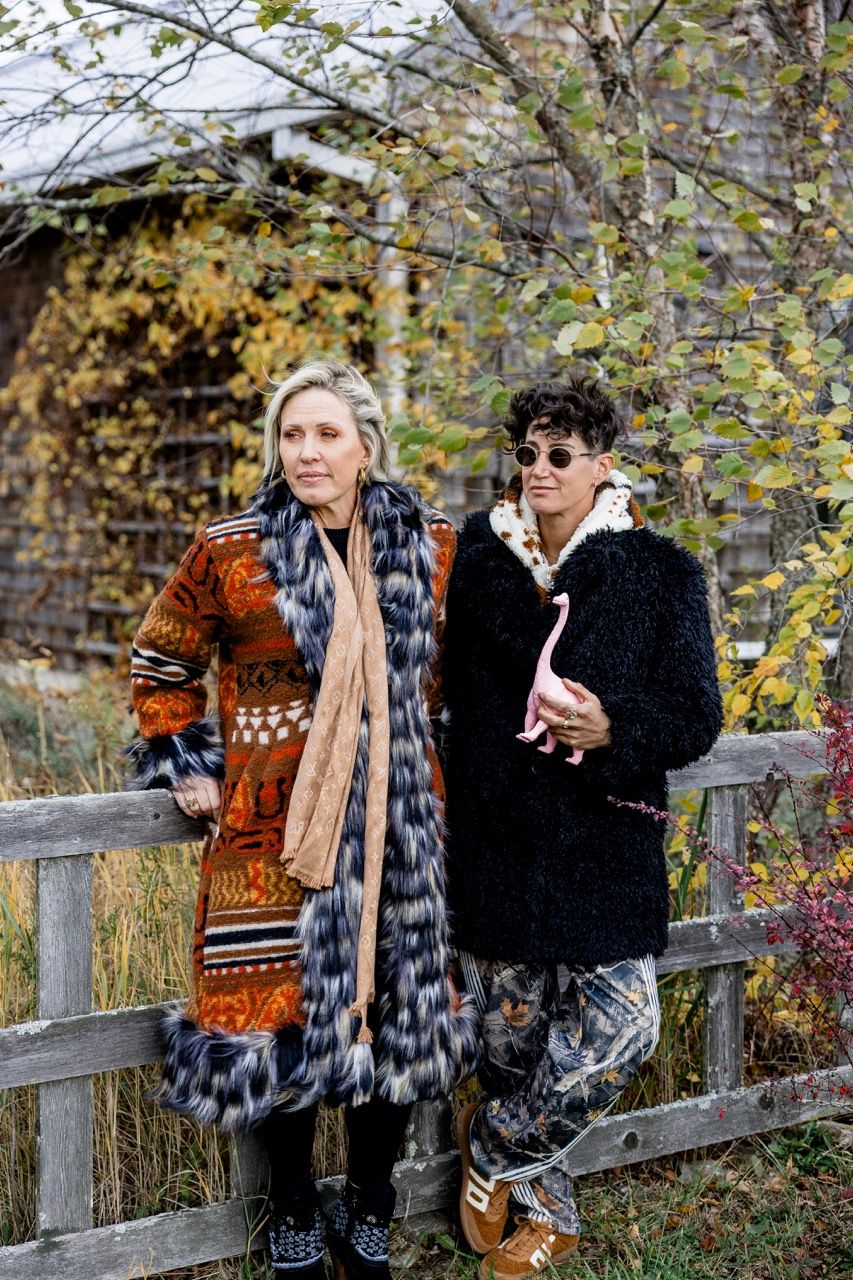Two stylishly dressed women pose by a rustic wooden fence surrounded by autumn foliage.